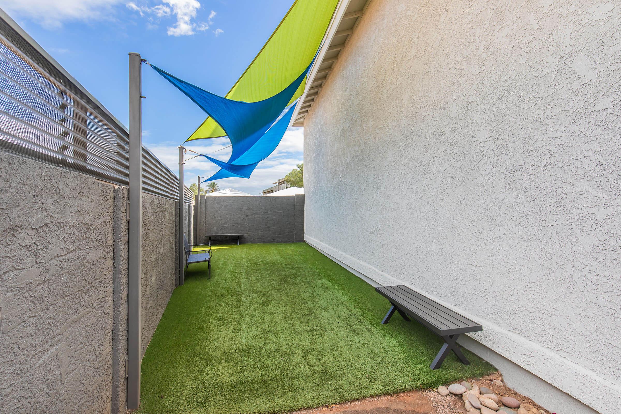 A narrow outdoor space featuring artificial grass, two black benches, and a colorful sail shade. The area is enclosed by gray walls and has clear blue skies overhead, creating a cozy environment for relaxation or leisure activities.