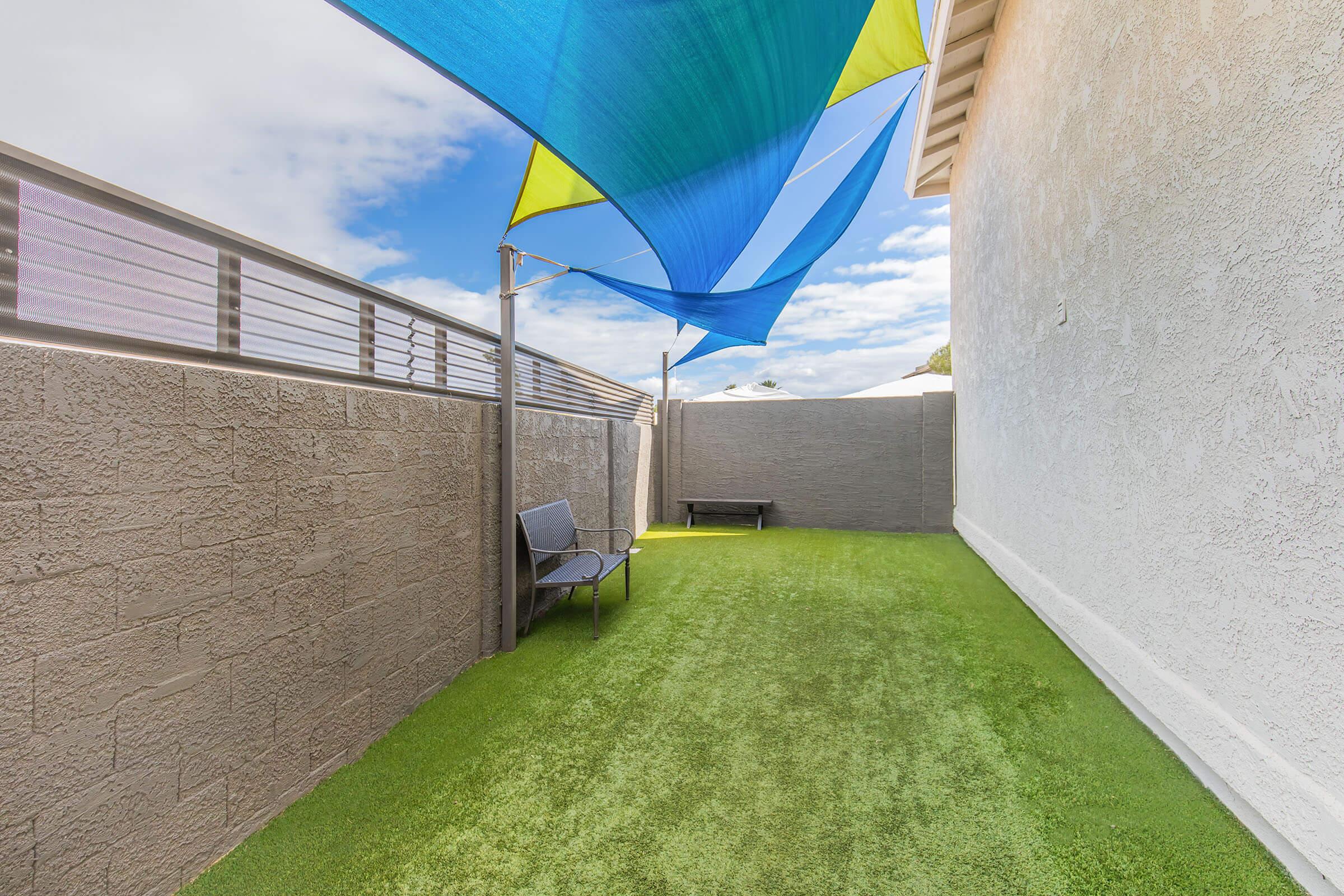 A small outdoor space featuring artificial turf, a single chair, and colorful shade sails providing protection from the sun. The area is enclosed by walls, with a clear blue sky visible above.