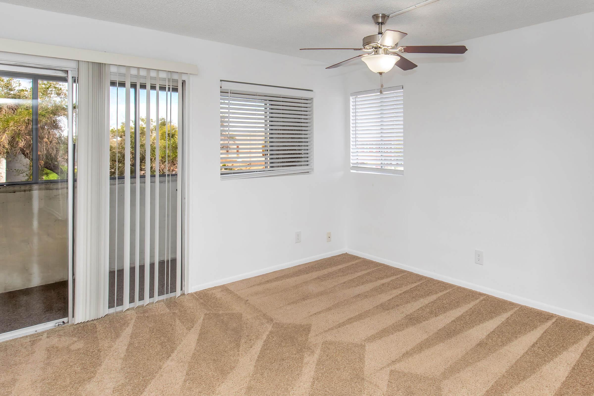 A bright, empty room featuring light beige carpeting, a ceiling fan, and large windows with white blinds. A sliding glass door leads to a balcony, allowing natural light to fill the space. The walls are painted white, creating an open and airy atmosphere.