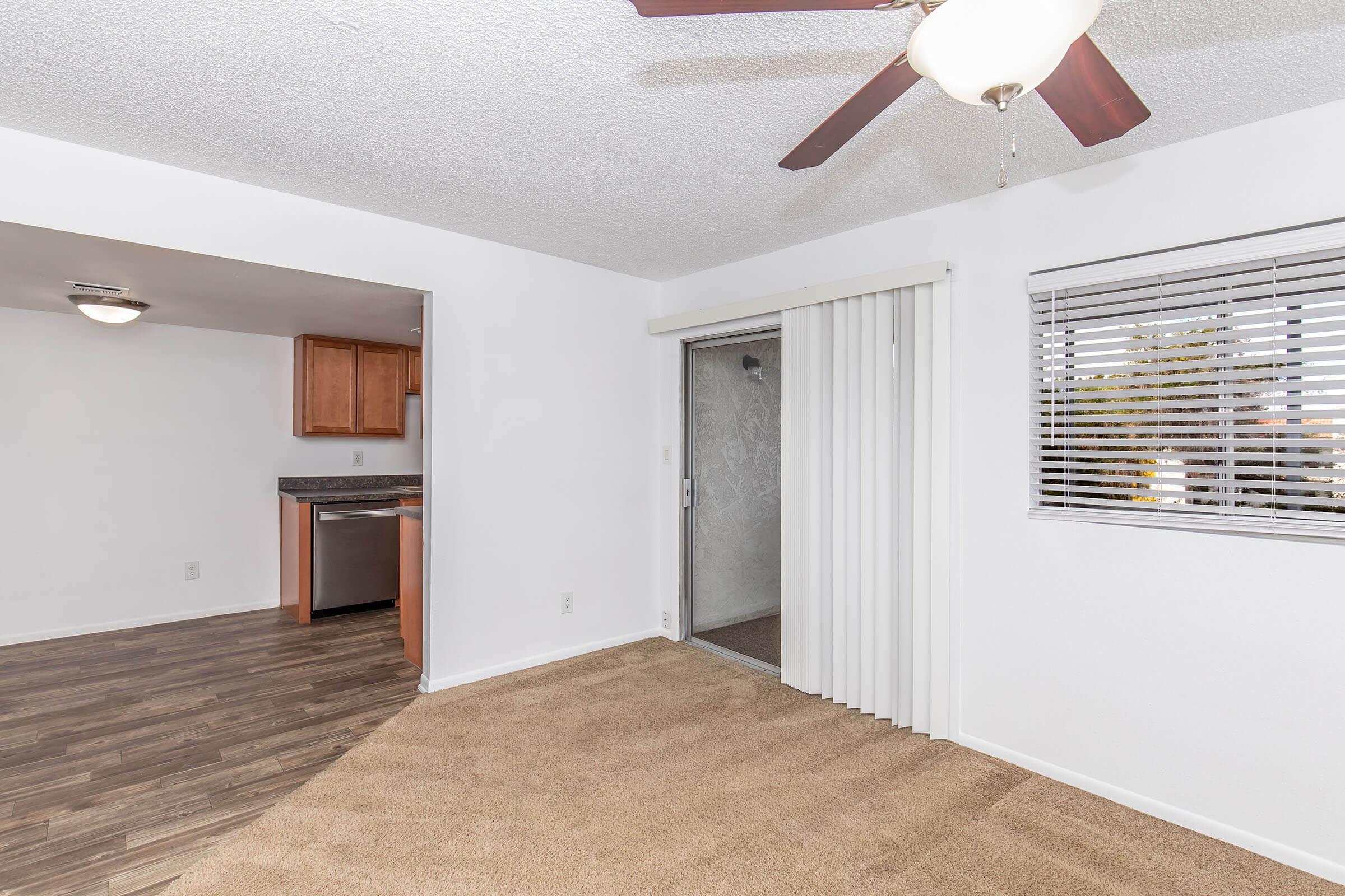 A cozy, well-lit living space featuring light-colored walls, carpet flooring, and a ceiling fan. To the left, there's an open kitchen area with wooden cabinetry and a countertop. A sliding door provides access to the outside, and there are window blinds allowing natural light to enter.