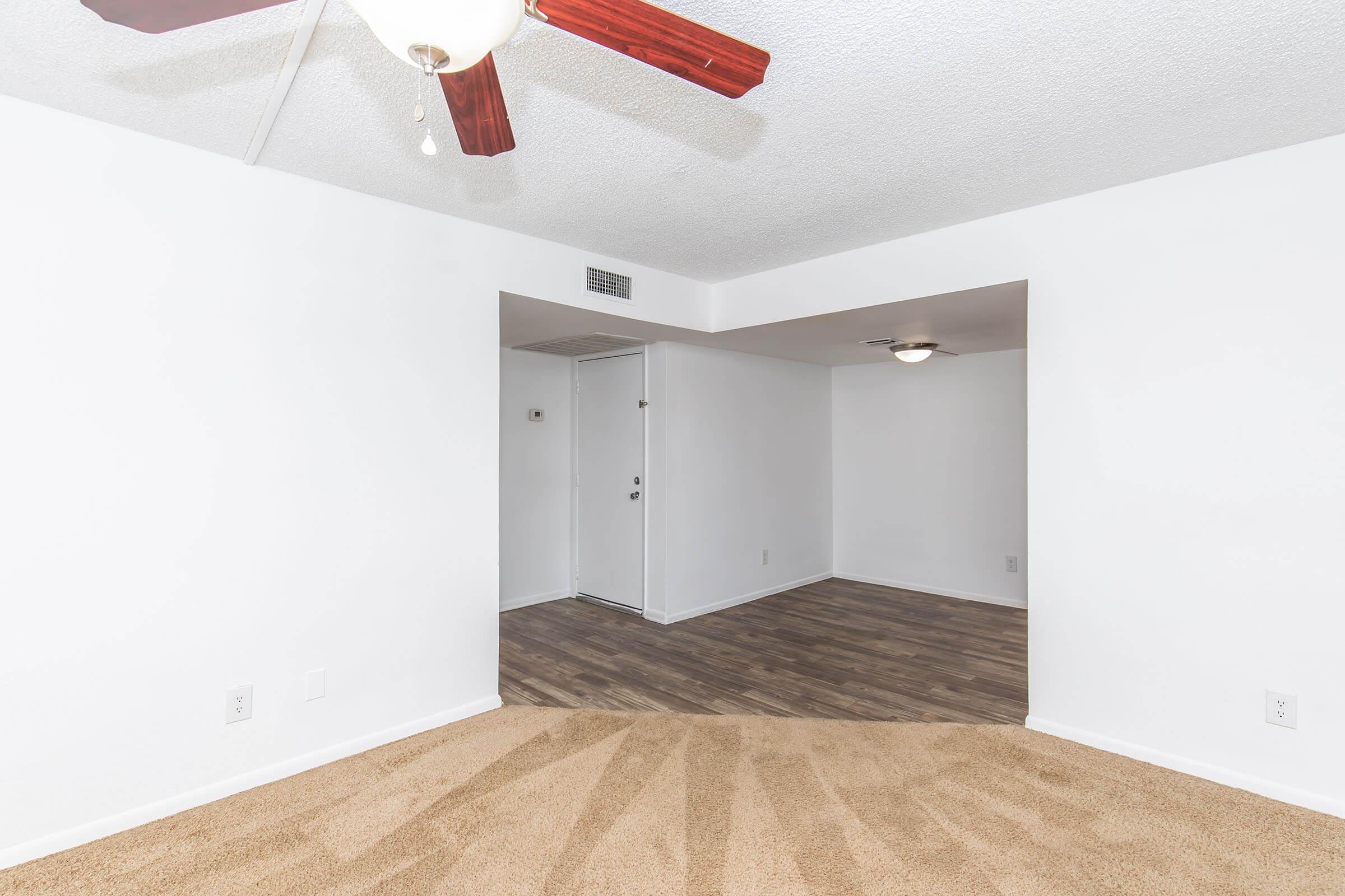 Interior view of a light-colored living space featuring a ceiling fan, beige carpet, and a wall with an opening leading to another room. A door can be seen in the background, indicating access to an entryway or hallway. The walls are painted white, and the flooring appears to be a dark wood laminate.