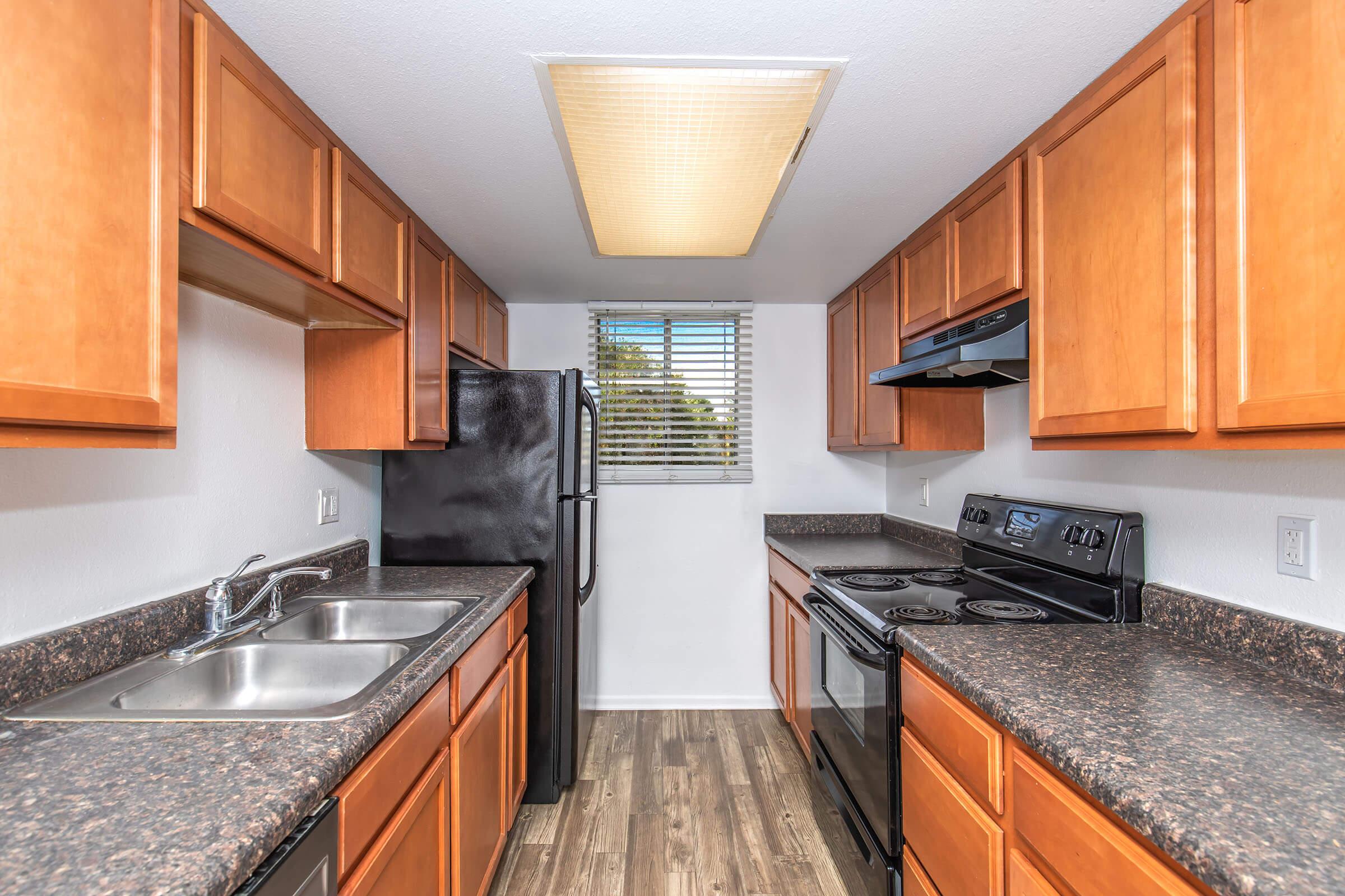 A modern kitchen with wooden cabinetry, stainless steel appliances, and a black refrigerator. The kitchen features a double sink, a window with blinds, and an overhead light. The countertops are dark, and the flooring is a light wood finish, creating a bright and functional space.