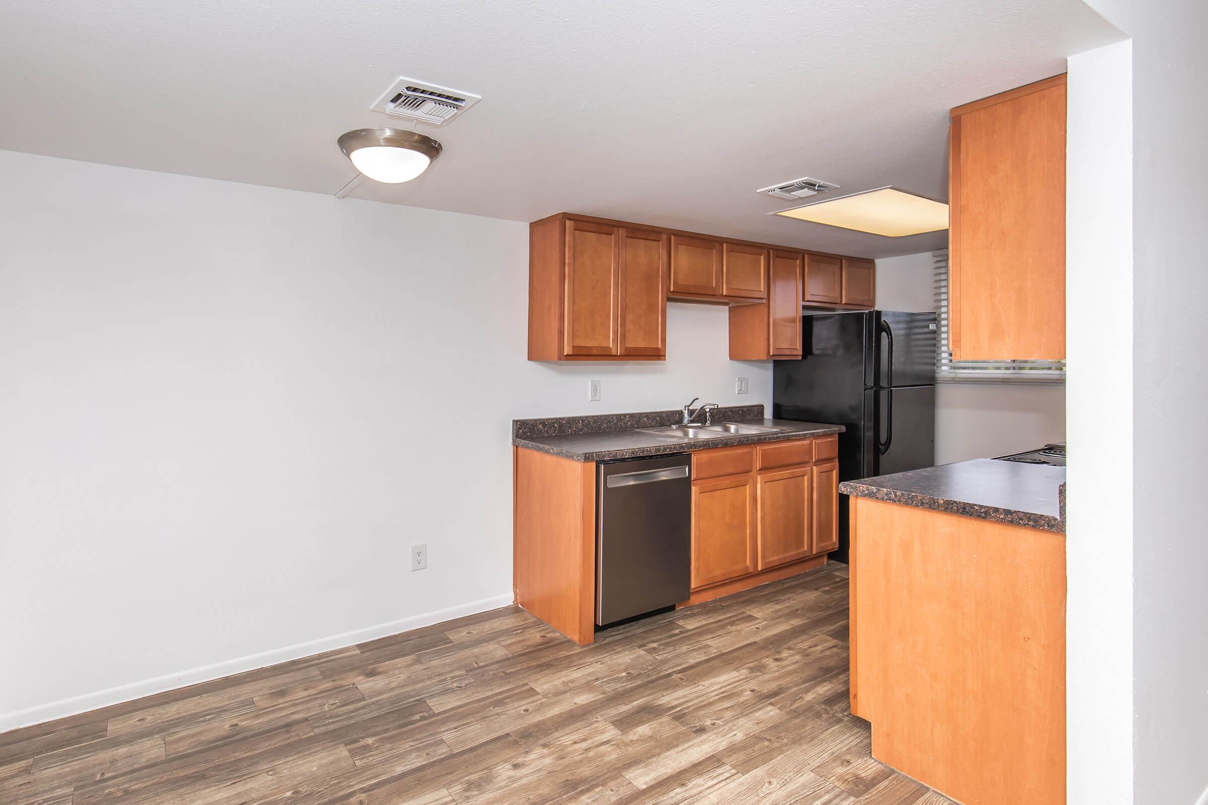A modern kitchen featuring wooden cabinets, stainless steel appliances including a dishwasher, and a dark countertop. The flooring is a warm wood finish, with ample lighting from a ceiling fixture and a window allowing natural light. The layout is open and spacious.