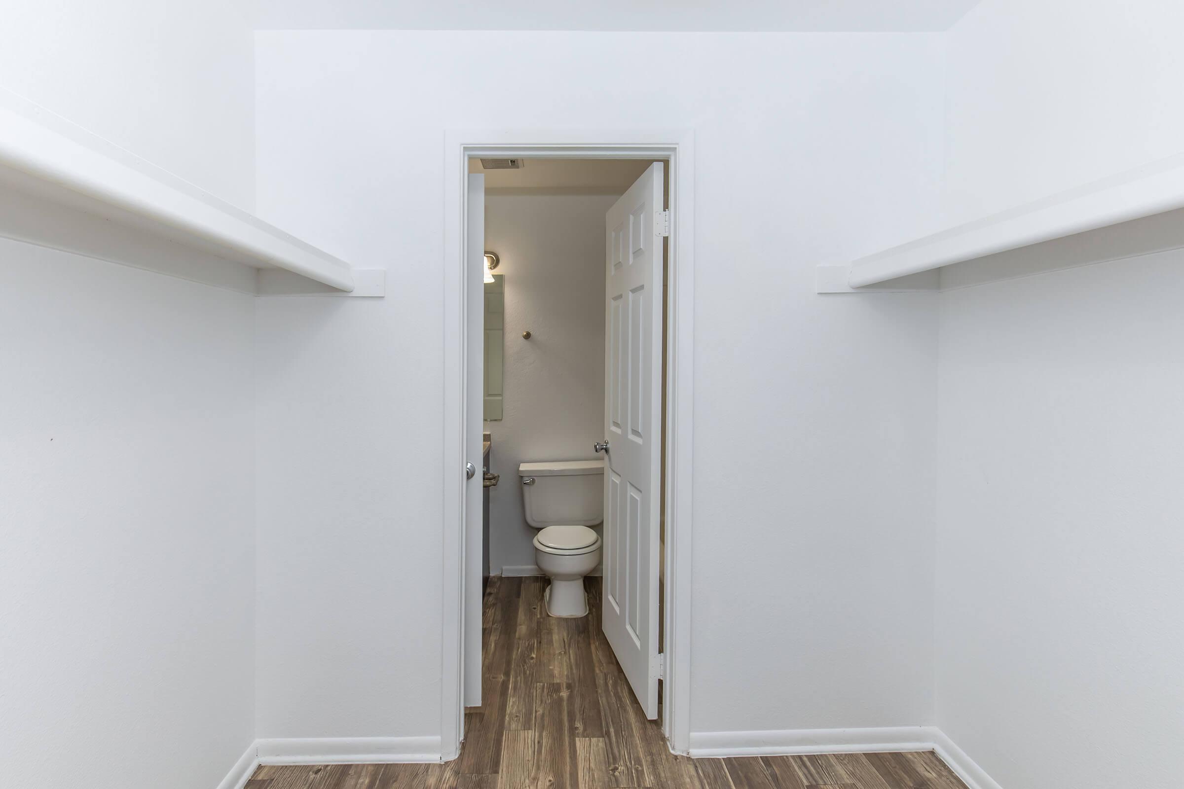 A spacious, empty room with white walls and hardwood flooring. On the left and right, there are shelves against the walls. In the background, a doorway leads to a bathroom featuring a toilet and a mirror. The room is well-lit and clean, suggesting a minimalist design.