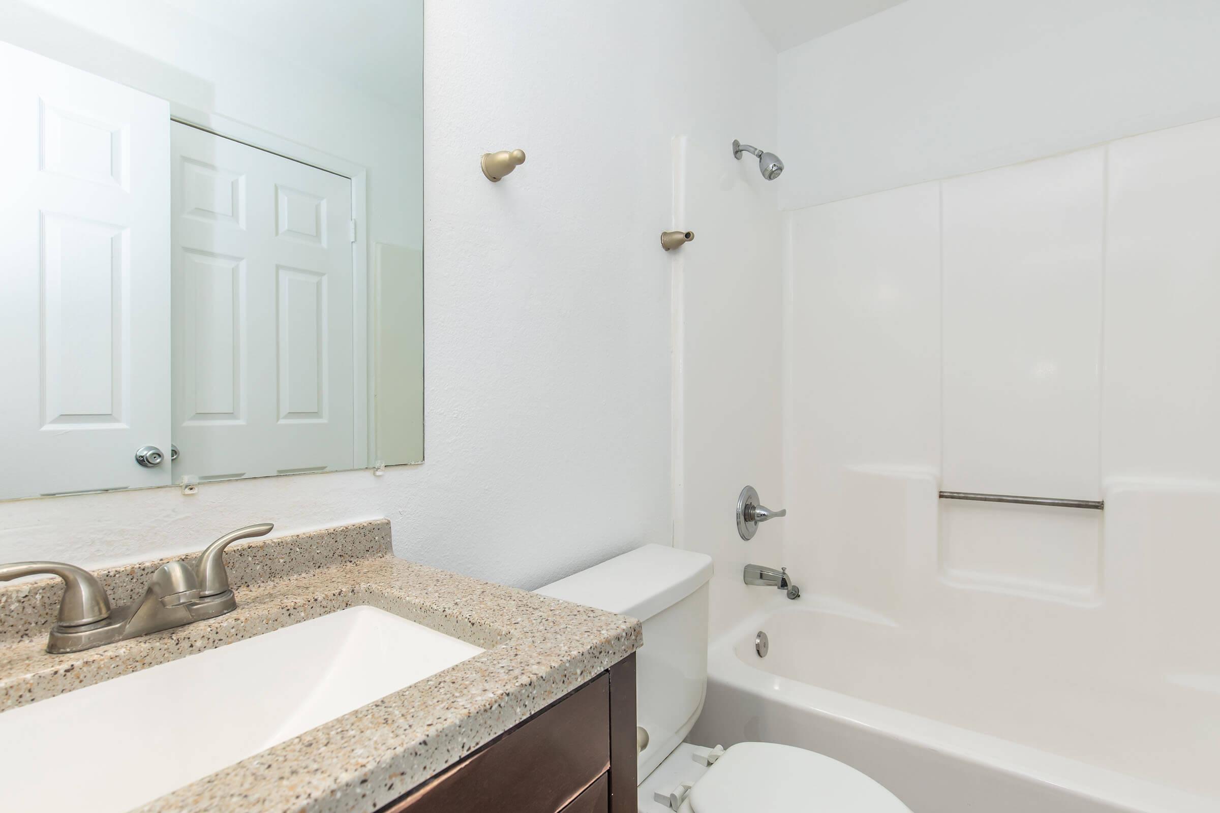 A clean and modern bathroom featuring a bathtub with a shower, a wall-mounted mirror, a granite countertop sink with dual faucets, and a toilet. The walls are white, and there's a door leading to another room. The overall aesthetic is bright and spacious.