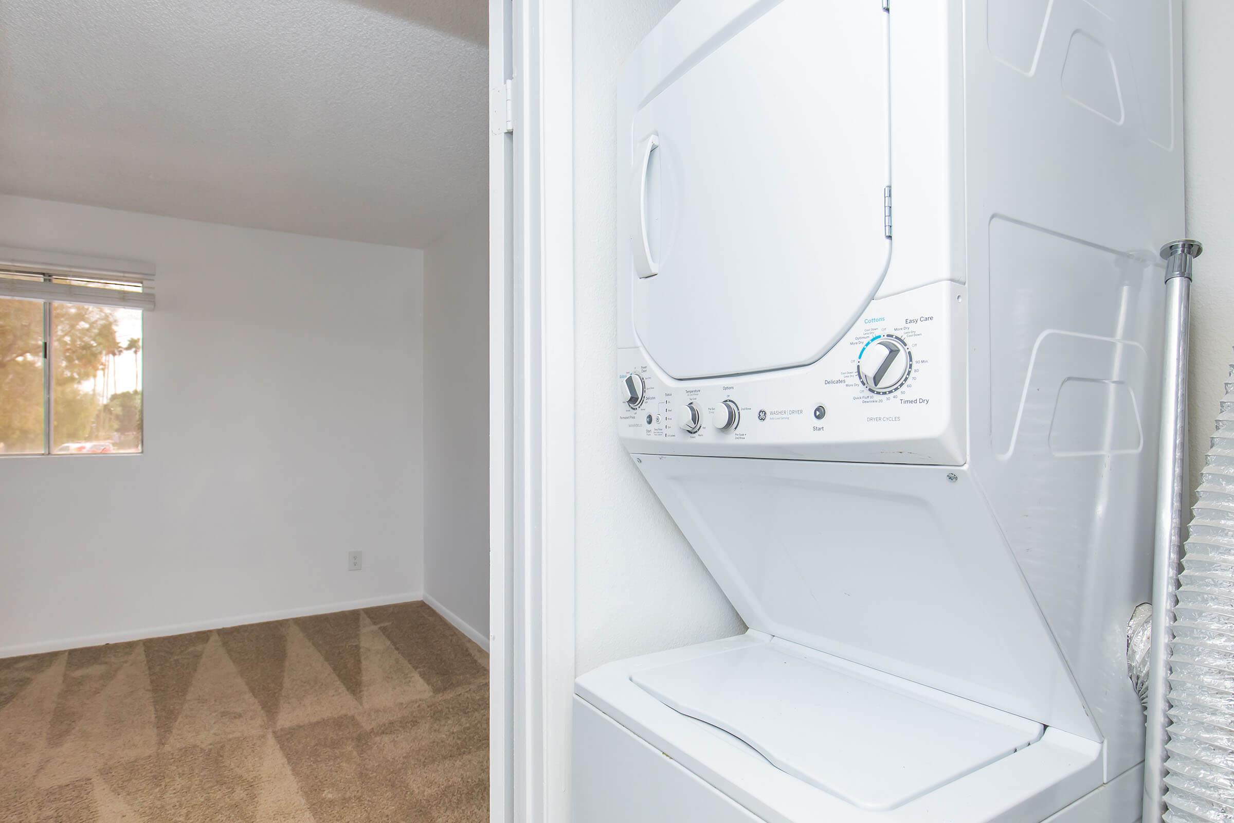 A stacked washer and dryer unit located in a laundry area with a window providing natural light. The adjoining room features beige carpet and white walls, creating a clean and open living space.