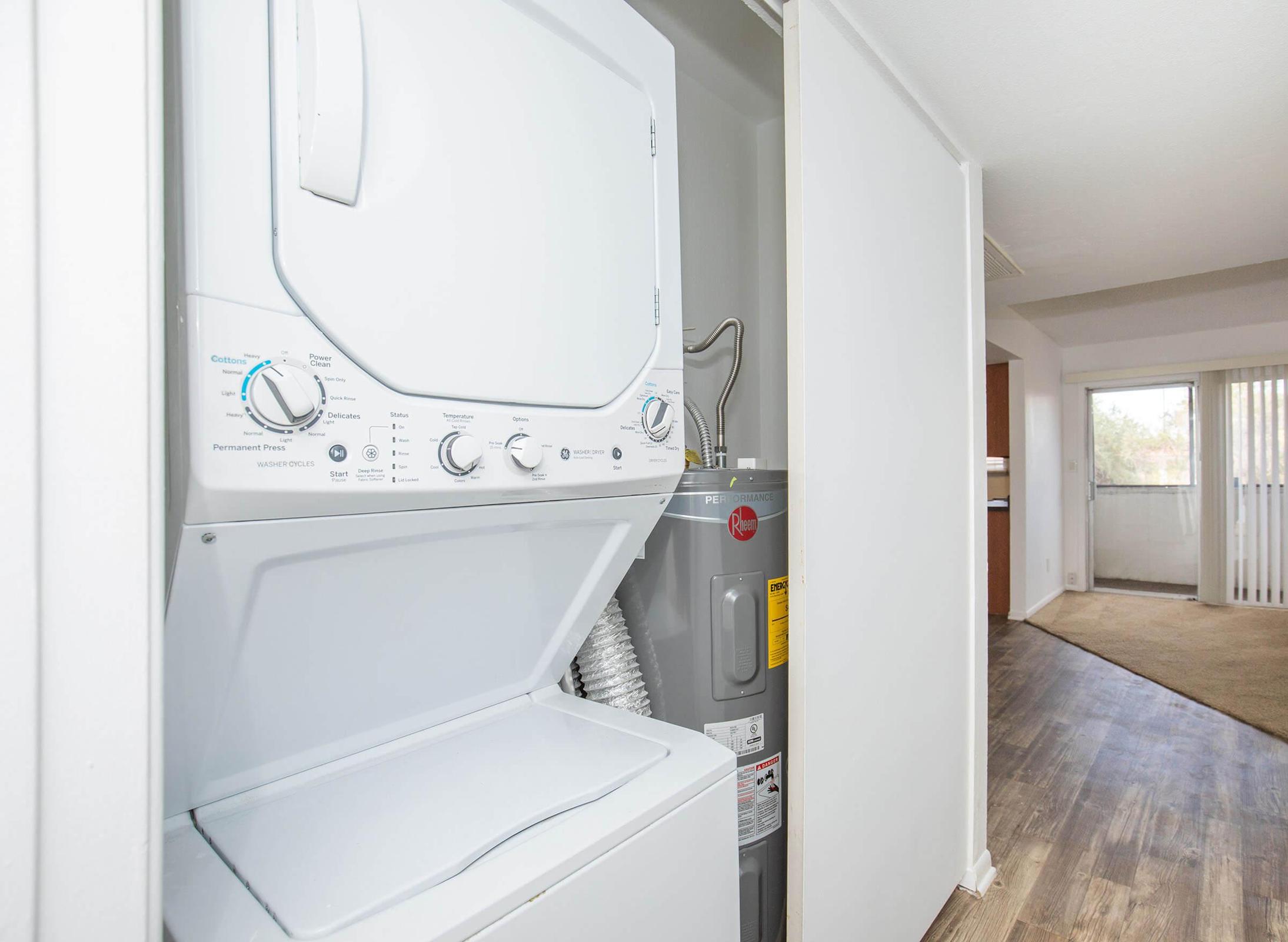 A stacked washer and dryer set is shown in a laundry area, with a water heater visible nearby. The space appears clean and organized, featuring light-colored walls and a hardwood floor, leading into a more spacious room with natural light.