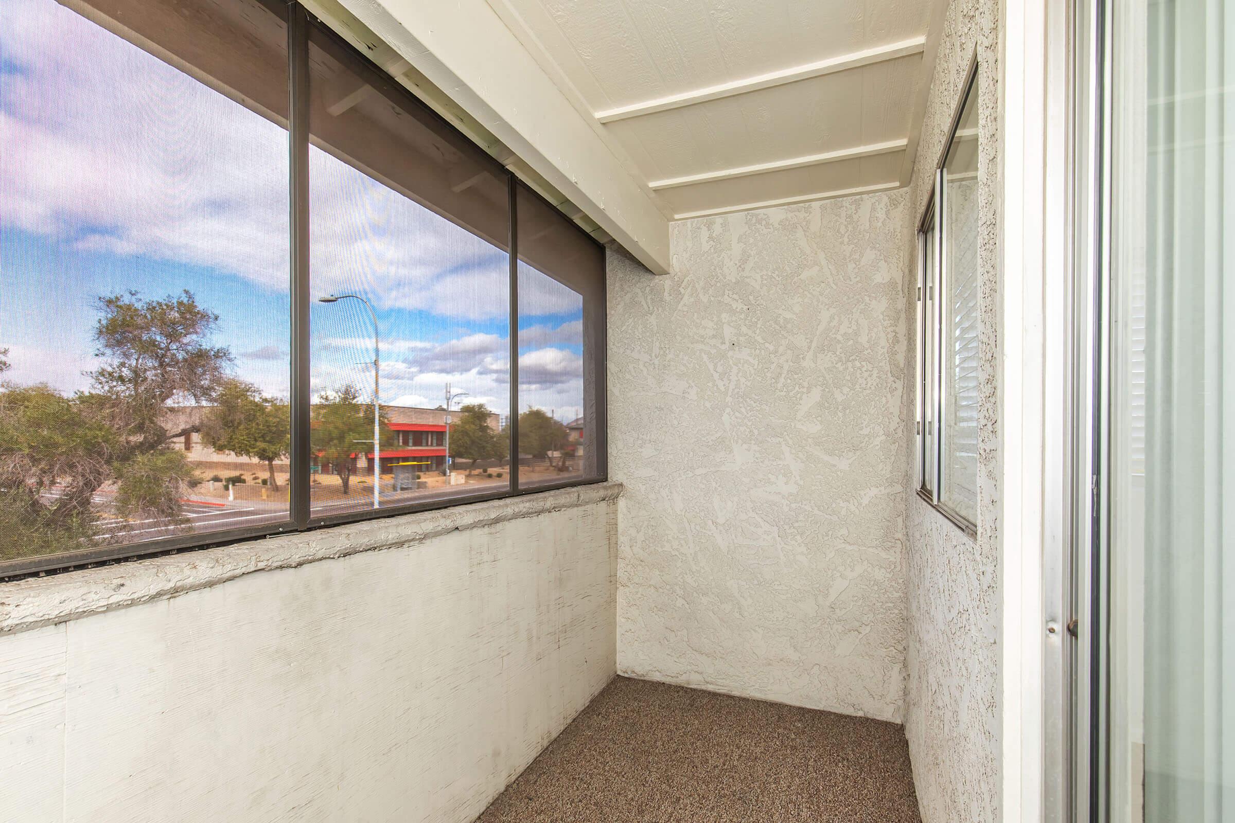 A small balcony with a textured wall, featuring large screened windows that allow natural light. There's a view of the outside with trees and buildings in the distance under a partly cloudy sky. The floor is covered in carpet or a similar material.