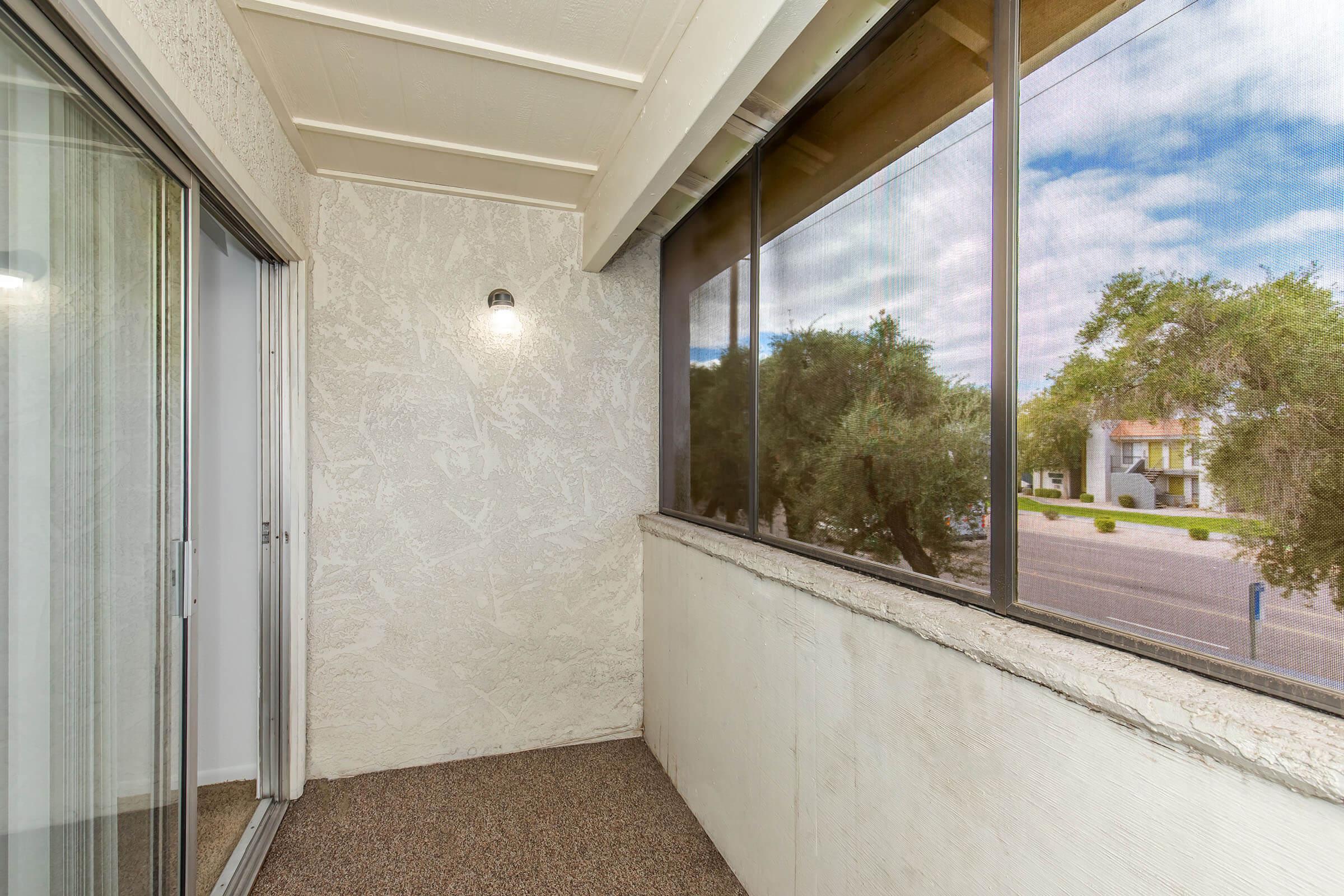 A small balcony with a textured wall and sliding glass doors. The space features a light fixture on the wall and a view of trees and a street outside. The floor is carpeted, and the overall ambiance is bright and airy with natural light filtering through the windows.