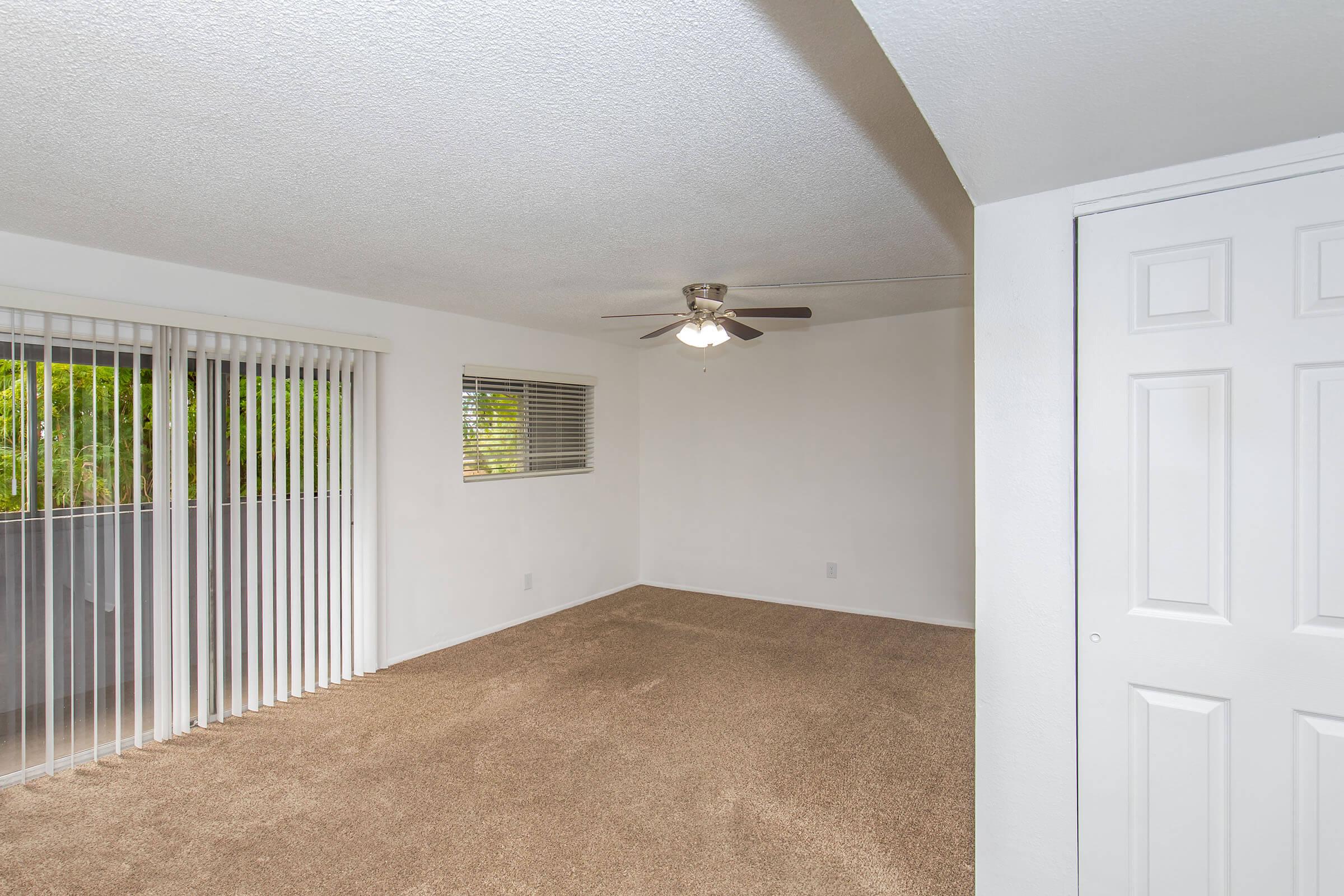 A spacious living area featuring light beige carpeting, a ceiling fan, and large sliding glass doors with vertical blinds that provide access to a balcony. The walls are painted in a neutral tone, and there's a closet with white double doors on the right side of the image.