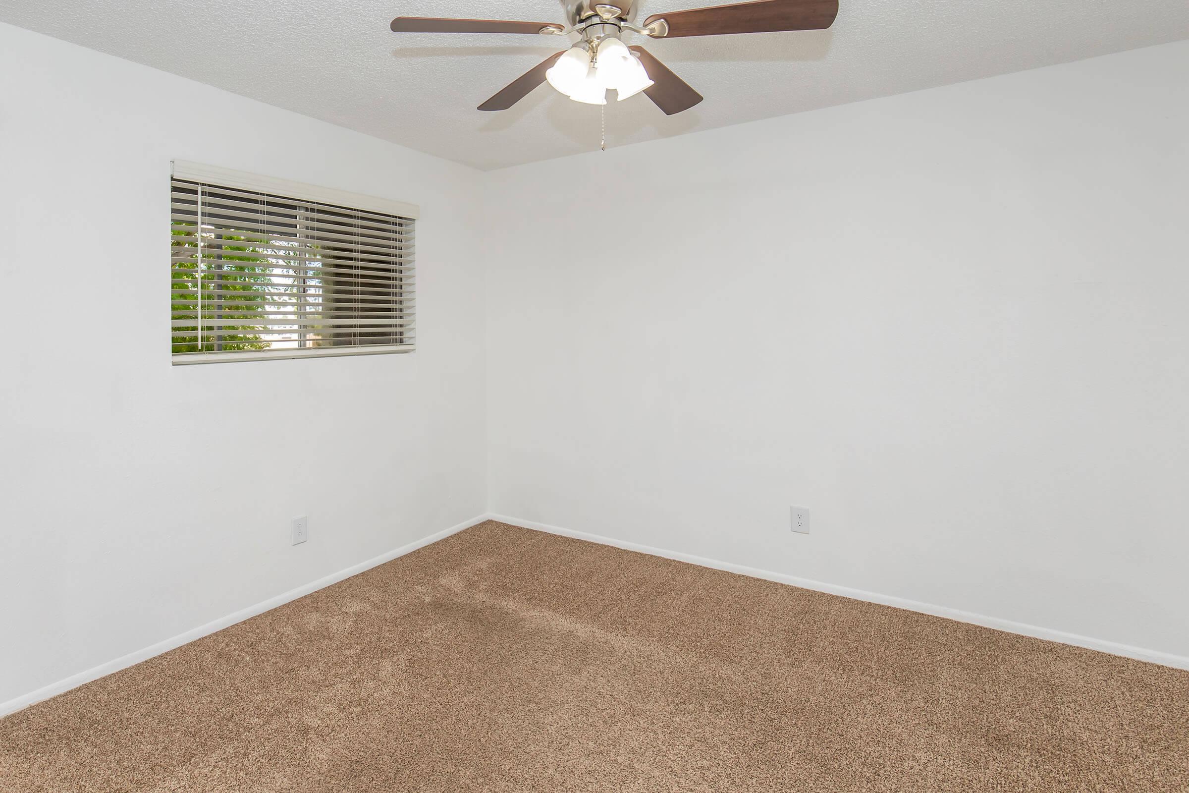 A bright and empty room with light beige carpet and white walls. There is a window with blinds letting in natural light, and a ceiling fan with three lights hanging in the center of the room. The space is clean and uncluttered, ideal for personal decoration.
