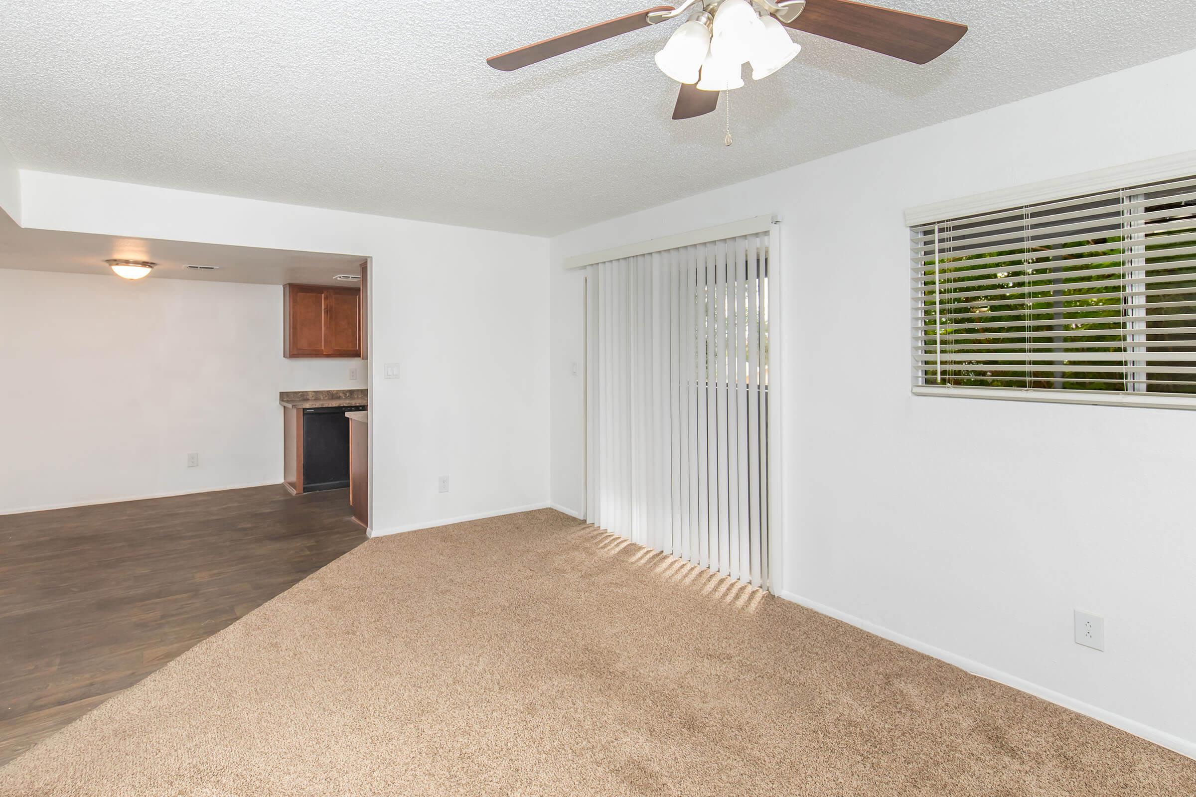 Spacious living area featuring a ceiling fan, beige carpeting, and large windows with vertical blinds. A doorway leads to a kitchen with wooden cabinetry. The room is brightly lit and has a neutral color scheme, creating a welcoming and open atmosphere.