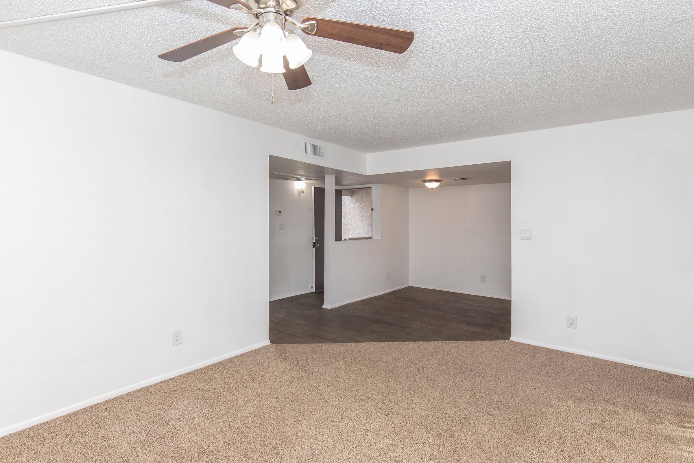 A spacious living area featuring light-colored walls, carpeted flooring, and a ceiling fan. A doorway leads to another room with a window and dark flooring, creating a cozy, inviting atmosphere. Natural light brightens the space, enhancing its openness.