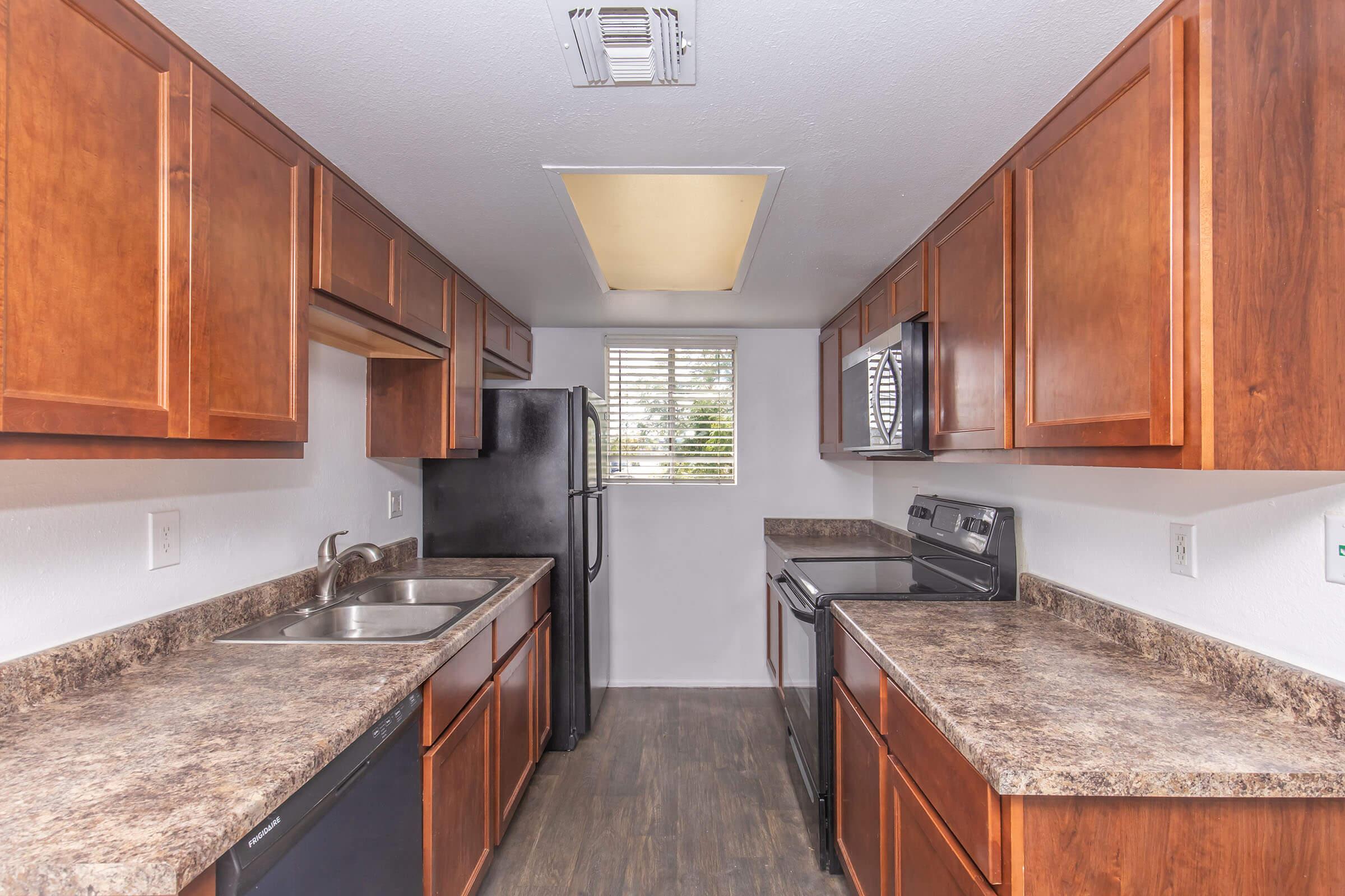 Modern kitchen featuring dark wood cabinetry, stainless steel appliances, a dual sink, and granite countertops. Large window provides natural light, and the layout includes a refrigerator, microwave, and oven. The flooring is a dark laminate, enhancing the contemporary look.
