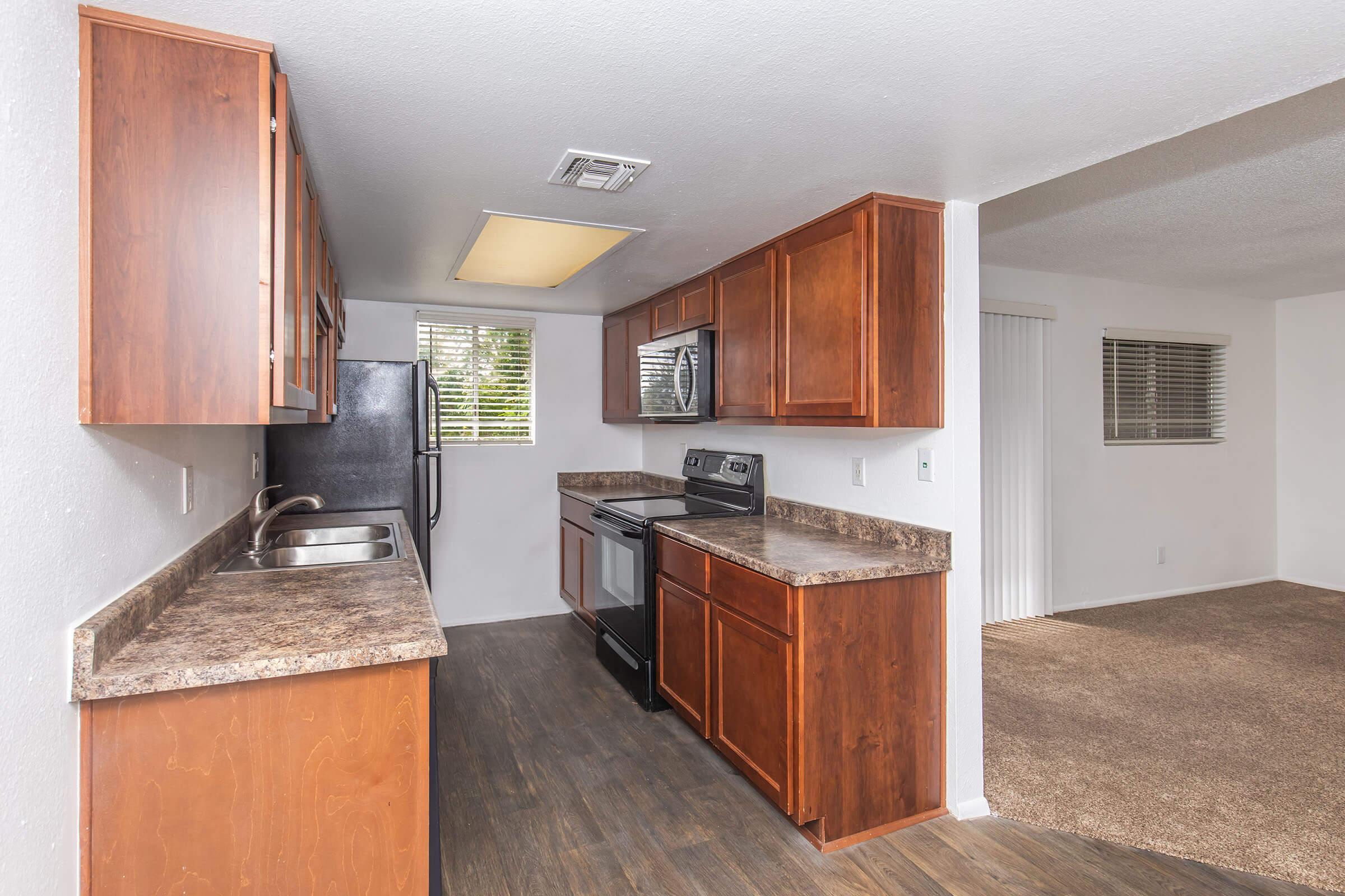 Interior view of a modern kitchen featuring dark wooden cabinets, a stainless steel refrigerator, and an oven. The kitchen has a countertop, and a window providing natural light. Adjacent to the kitchen is a carpeted living area, illuminated by soft light.