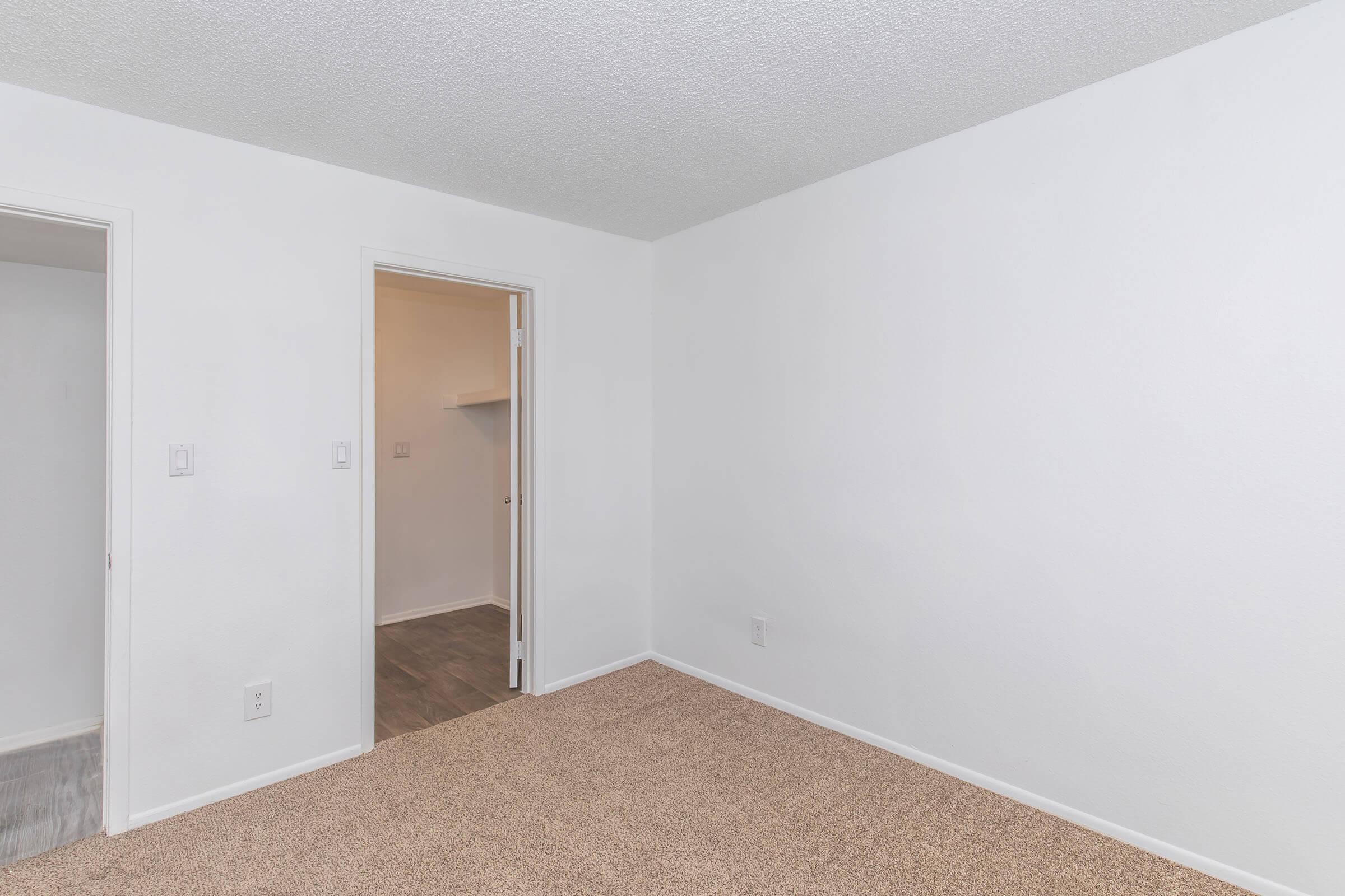 A view of an empty room with white walls and a carpeted floor. There are two doors: one leading to another room and another possibly leading to a closet. The lighting is neutral, and the space appears clean and minimalist.