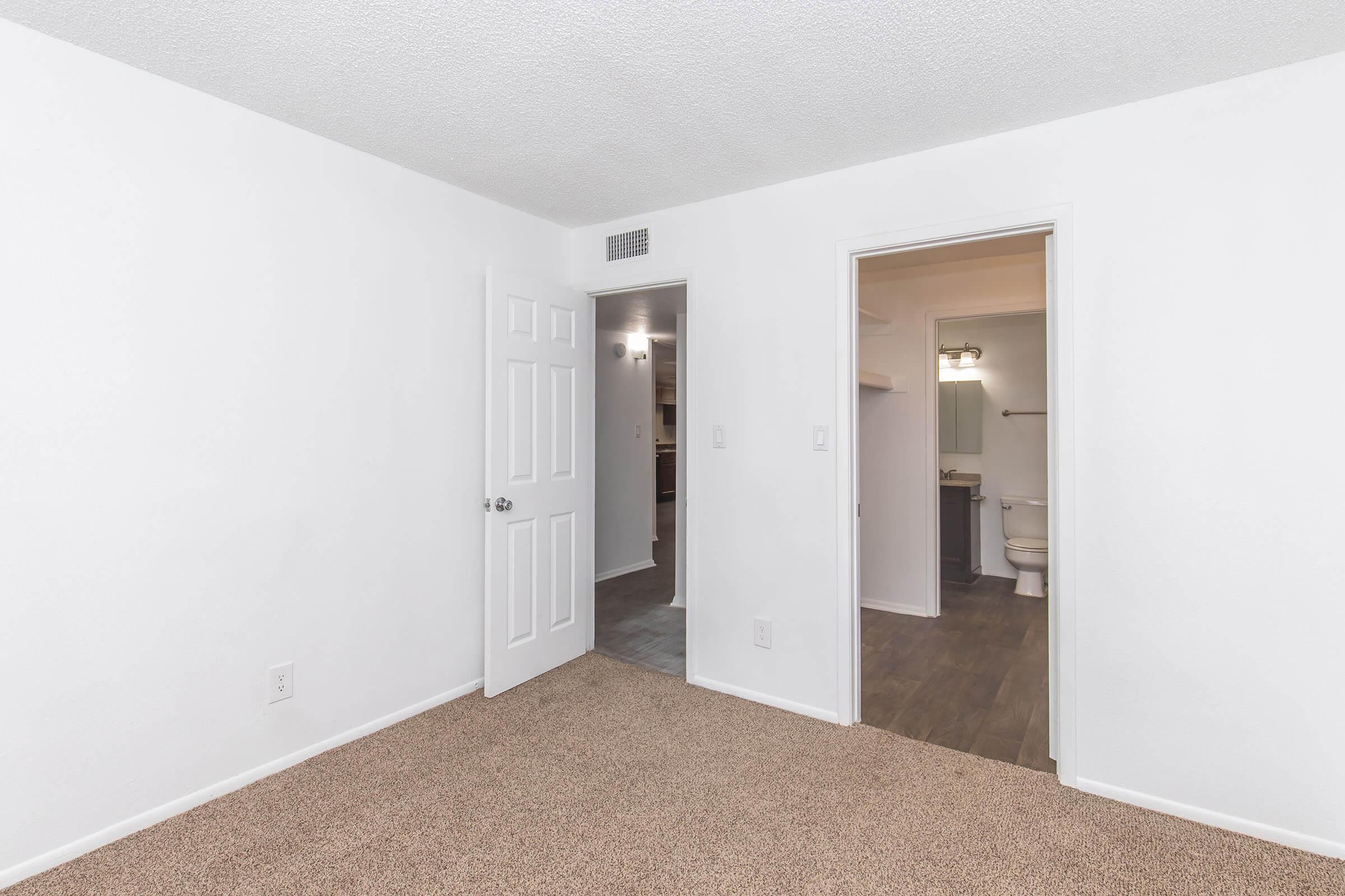A small, well-lit room with beige carpet and white walls. There are two doors: one leading to a bathroom and the other to another room or hallway. The space is simple and unfurnished, creating a clean and open feel.
