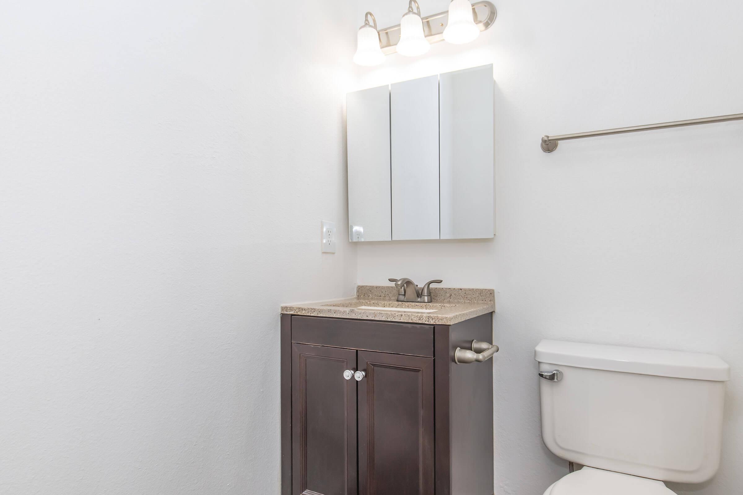 A clean, modern bathroom featuring a countertop sink with a dark wood vanity, a mirror above, and a toilet. The walls are painted white, and there is a chrome towel bar. Soft lighting from overhead fixtures illuminates the space.