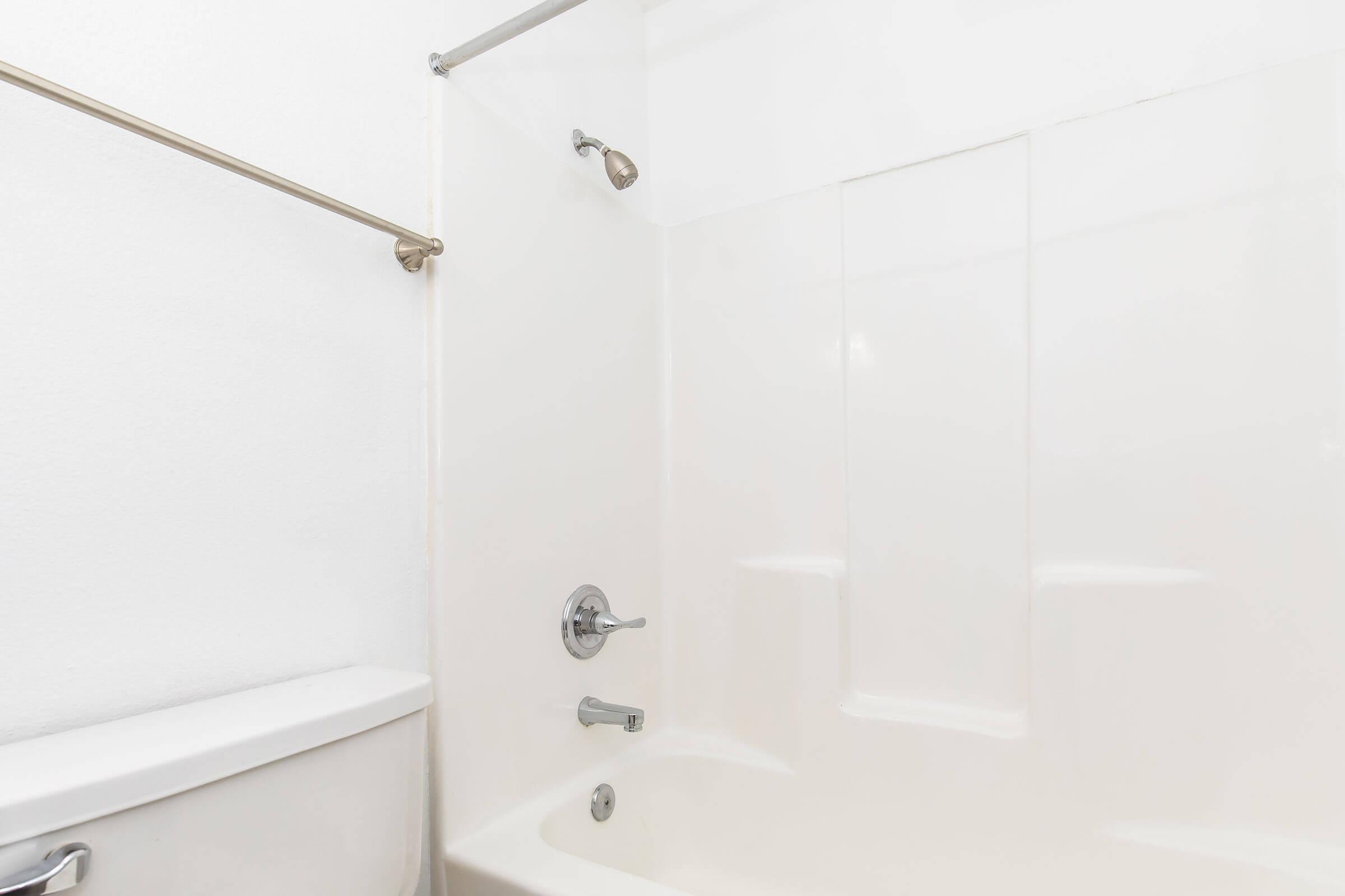 A clean bathroom featuring a white bathtub and shower with a chrome faucet and showerhead. The wall is a plain white, and there is a metallic grab bar alongside the bathtub. Toilet visible to the left. The overall aesthetic is simple and modern.