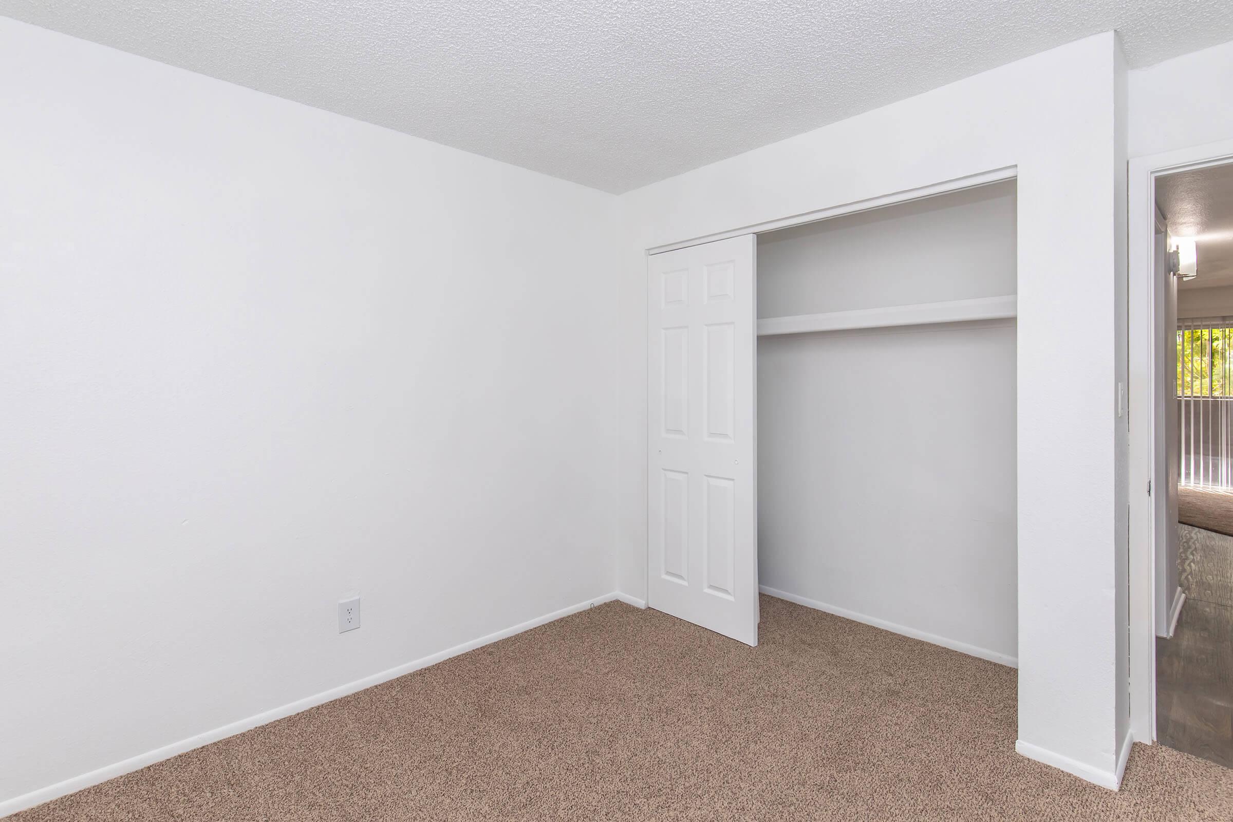 Empty room with white walls and a light brown carpet. There is a closet with sliding doors on one side, featuring a shelf inside. The room appears well-lit and spacious.