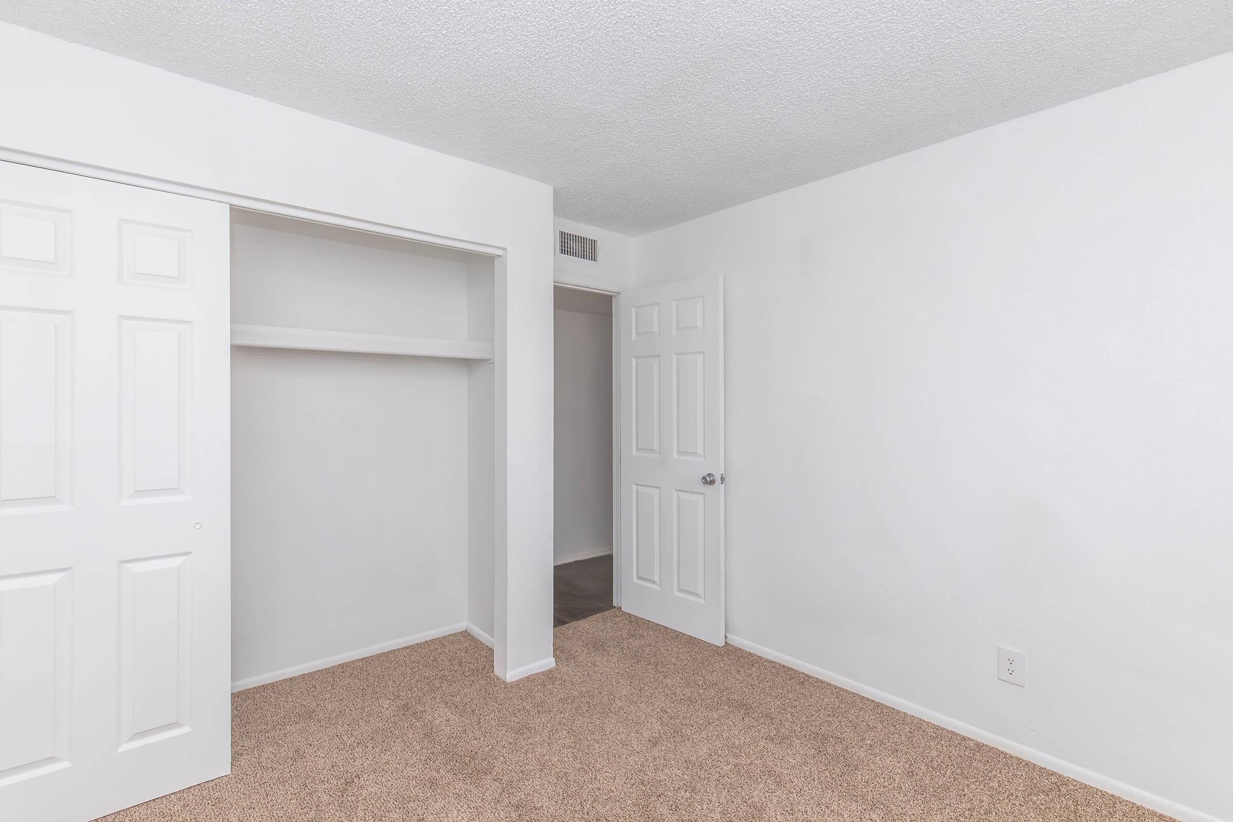 Empty room featuring light-colored walls, beige carpet flooring, and a closet with sliding doors. A doorway visible on the right leads to another space, while the overall atmosphere appears bright and clean.