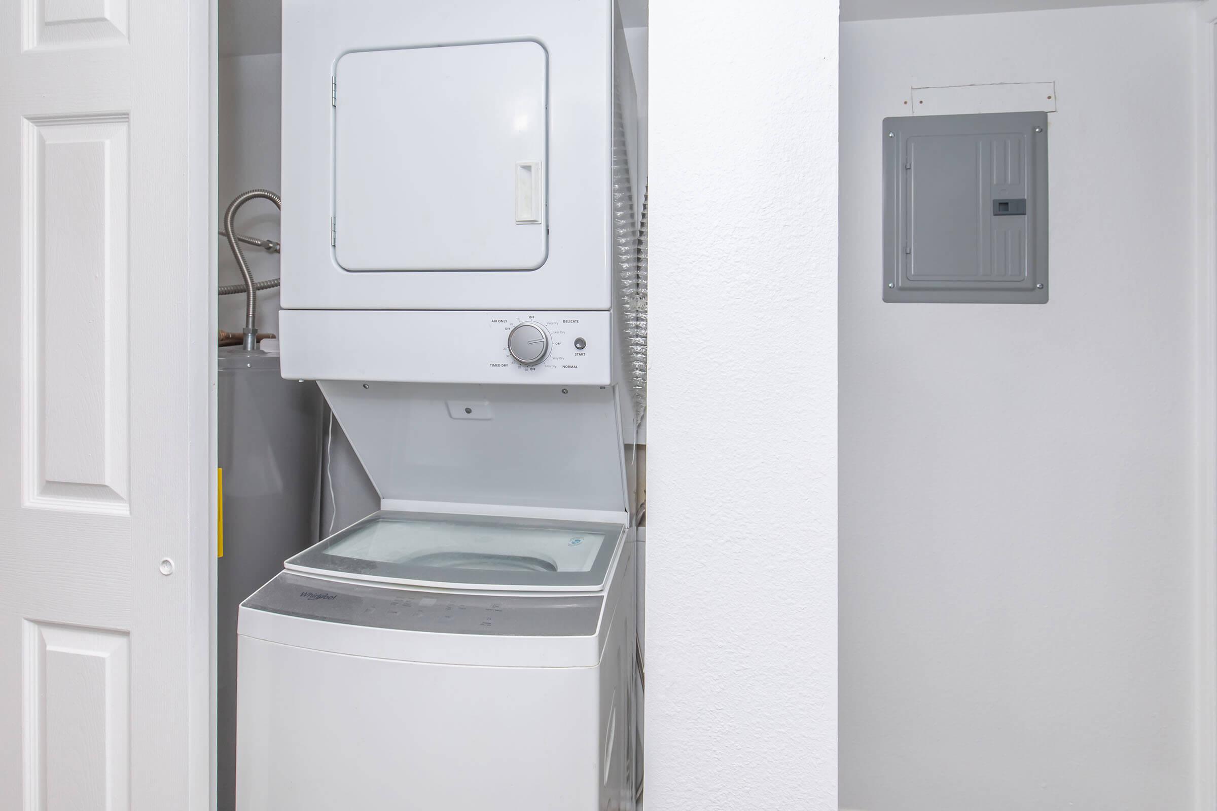 A compact laundry area featuring a stacked washer and dryer unit against a clean white wall. To the right, there is an electrical panel, and to the left, a water heater is partially visible. The space is well-lit and organized, showcasing a functional design for laundry needs.