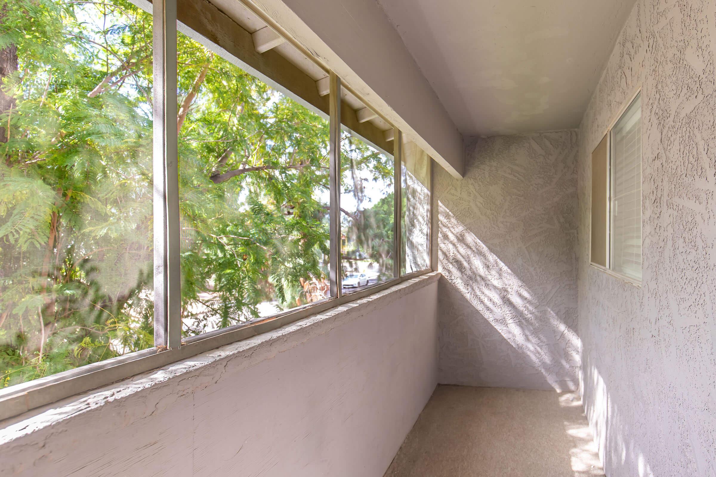 A bright, airy balcony with large windows allowing natural light to flood in. The view shows greenery from nearby trees, creating a peaceful and serene atmosphere. The walls are light-colored, and the floor is plain and unadorned, making the space feel open and inviting.