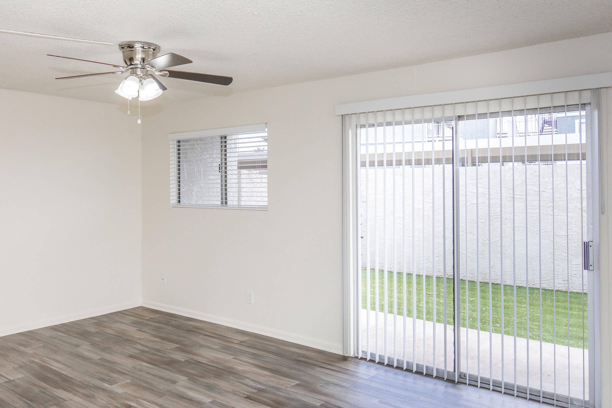 Interior view of a living room featuring gray wood-like flooring, a ceiling fan with light fixtures, and a sliding glass door with vertical blinds leading to a small outdoor area. A window with white blinds is visible on the left, allowing natural light into the room. The walls are painted a light color.