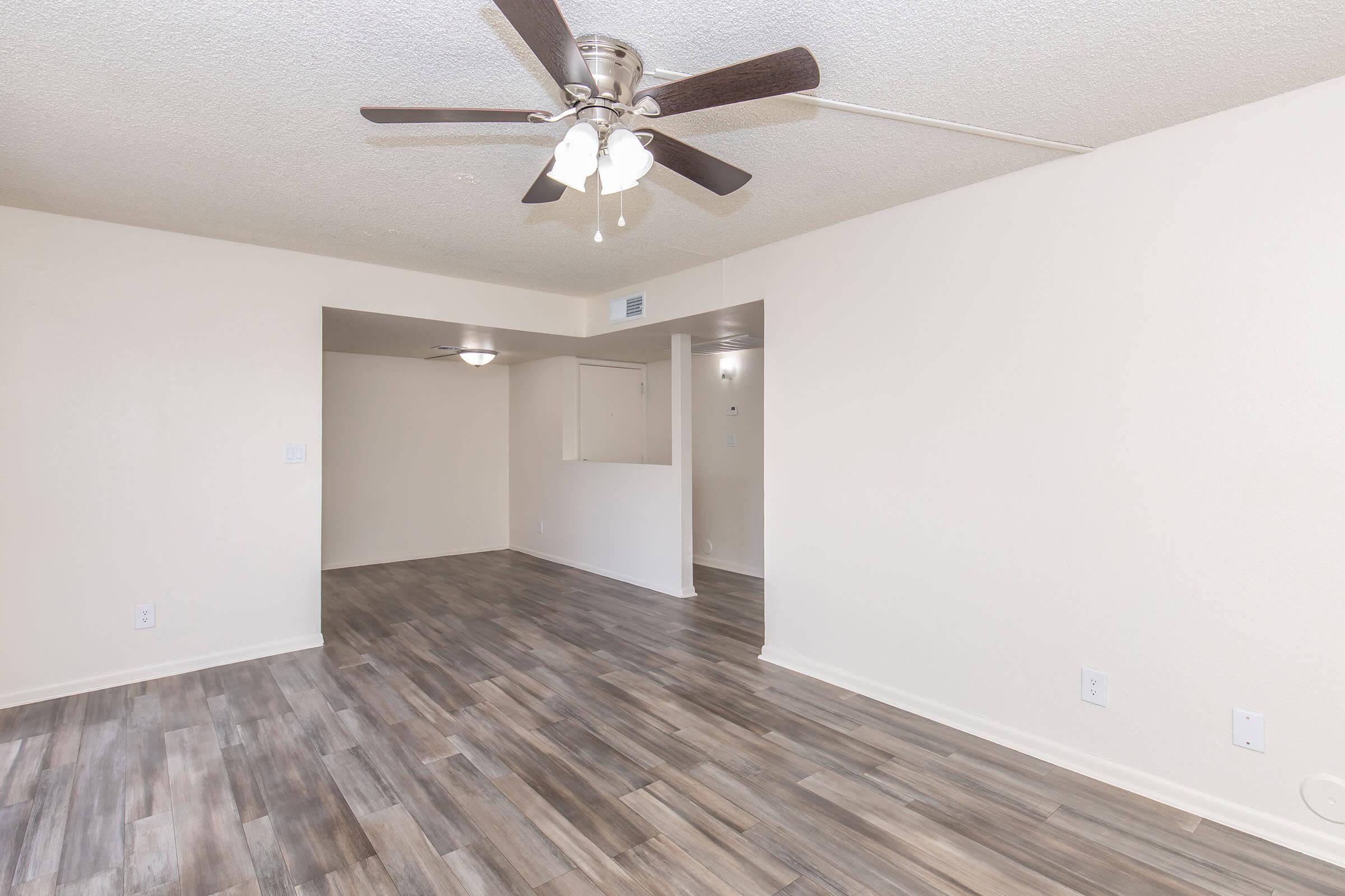 A spacious, empty living room featuring a light-colored ceiling and walls, wooden-like flooring, and a ceiling fan with three light bulbs. Two openings lead to adjacent rooms, creating a bright and airy atmosphere. The overall design is modern and minimalistic, suitable for various decor styles.
