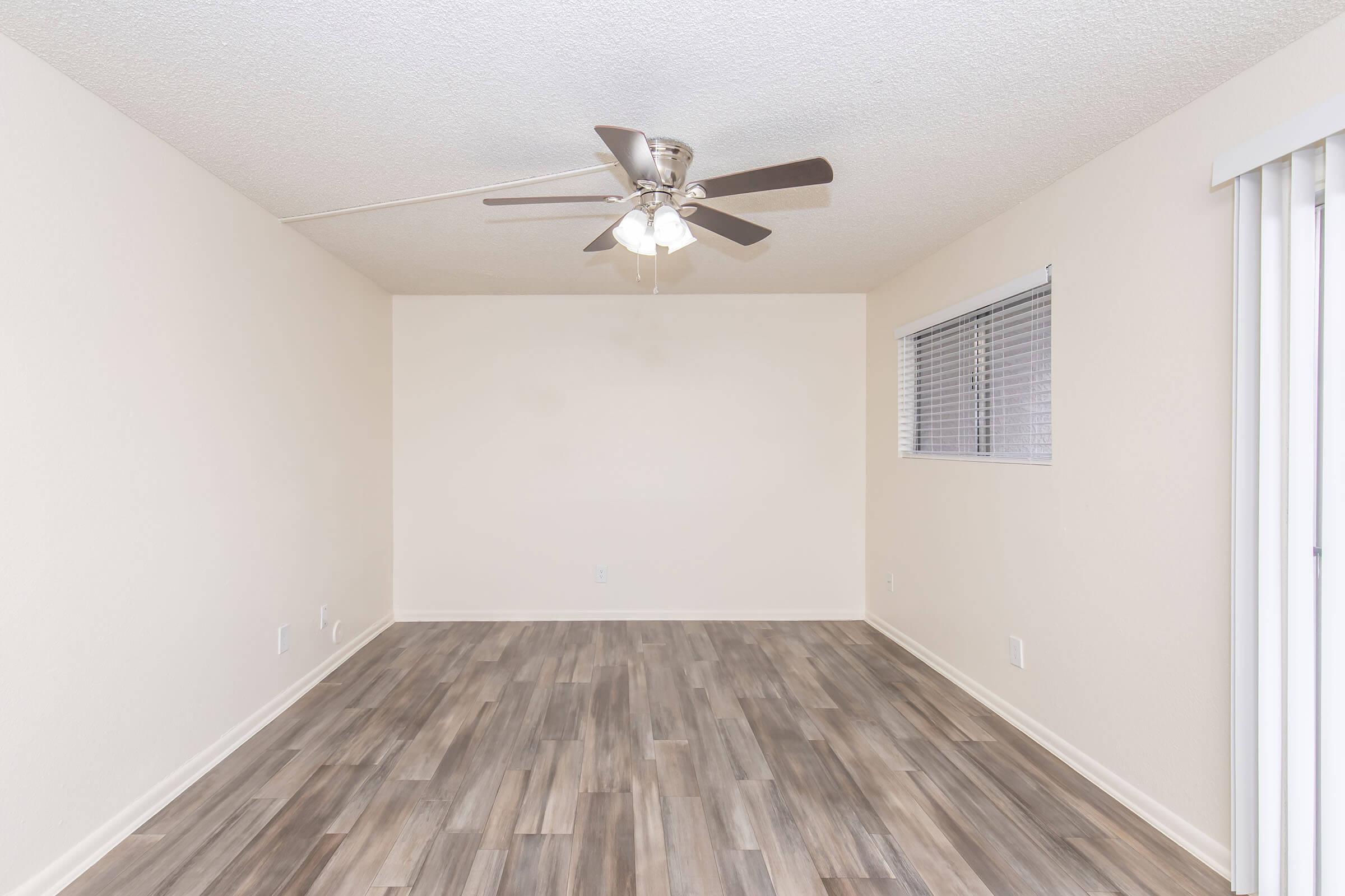 An empty room with light-colored walls and a ceiling fan. The floor has wood-like tiles, and there is a window with blinds on one wall. The space is well-lit, creating a bright and airy atmosphere.