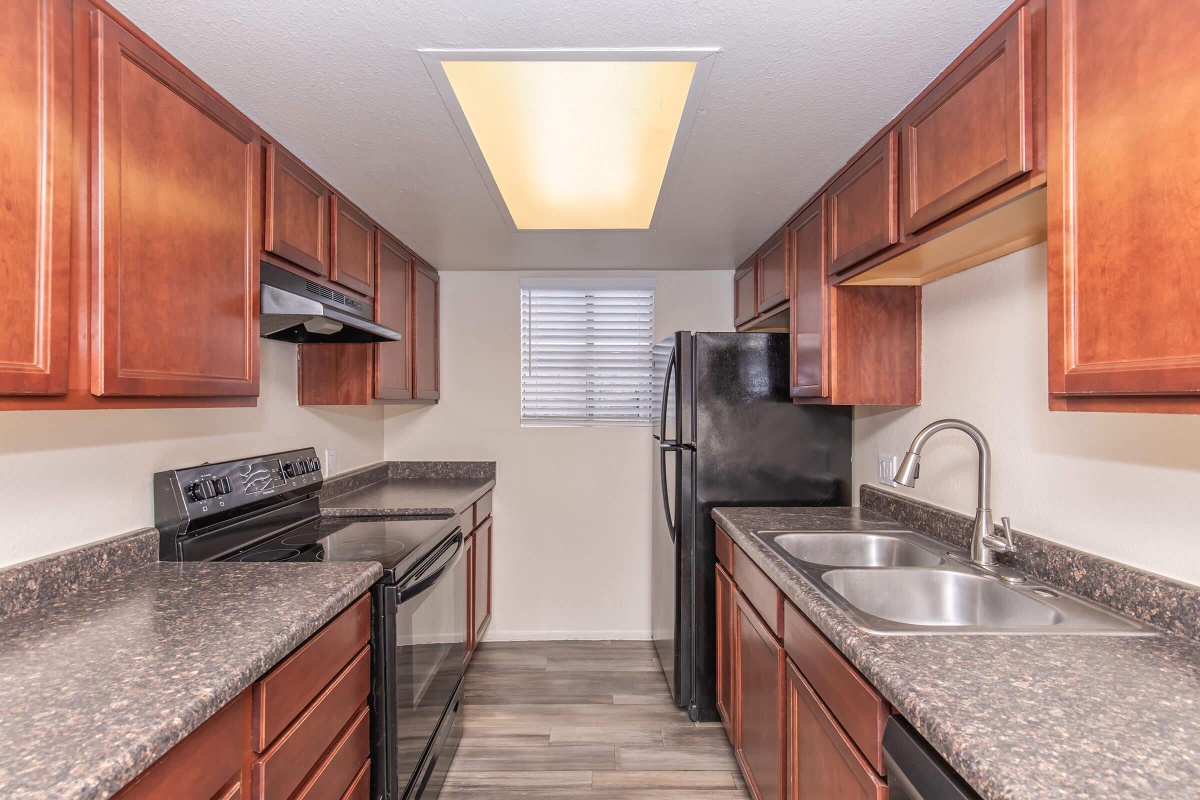A modern kitchen featuring wooden cabinets, granite countertops, stainless steel appliances, and dual sinks. The layout includes a black refrigerator and oven, with a window providing natural light above the sink area. The flooring is a sleek, dark laminate, contributing to the contemporary look.
