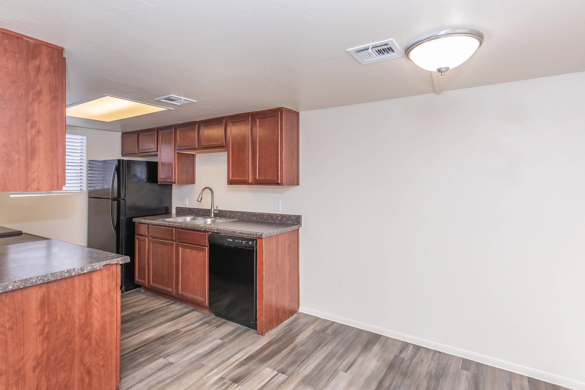 A modern kitchen featuring wooden cabinetry, a black refrigerator, a dishwasher, and a stainless steel sink. The space includes a countertop and an open layout with ample natural light from a nearby window, complemented by a ceiling light fixture. The flooring is a wood-like laminate.