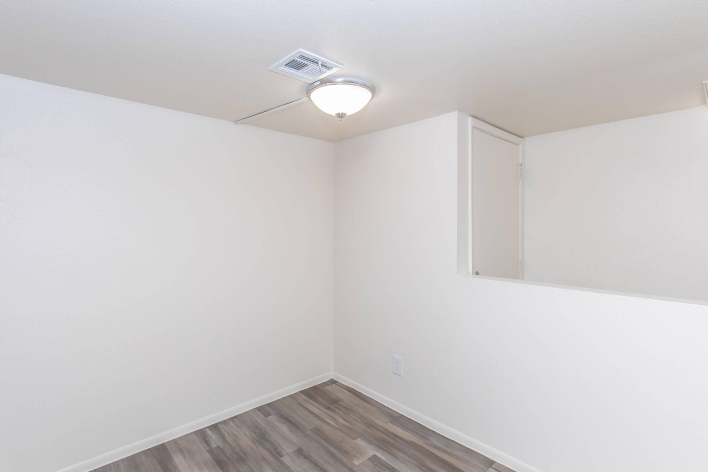 A corner of a minimally decorated room featuring light-colored walls, a ceiling light fixture, and laminate flooring. A small door is visible on one wall, and the overall appearance is bright and uncluttered, suggesting a modern living space.