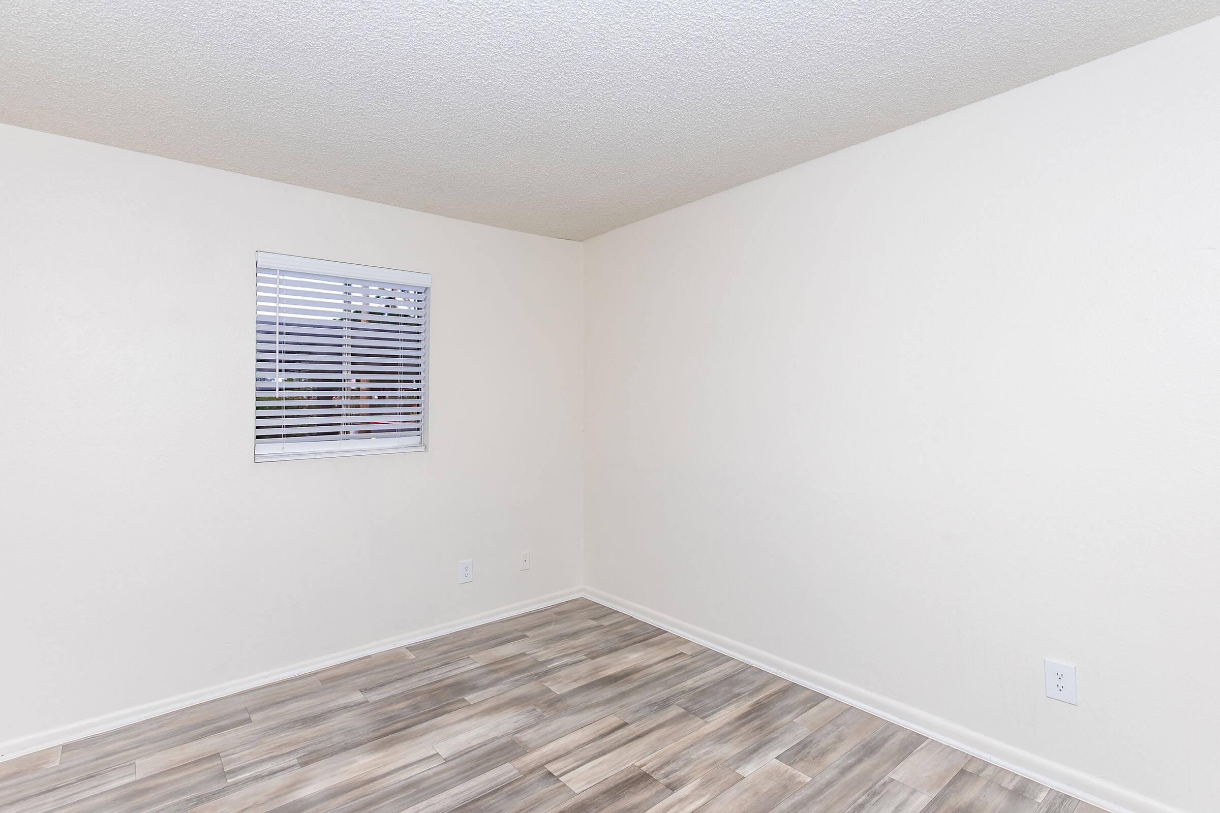 Empty room with light-colored walls and a window with white blinds. The flooring is wooden in appearance, featuring a blend of light and dark tones. The overall space is well-lit and uncluttered, creating a feeling of openness.