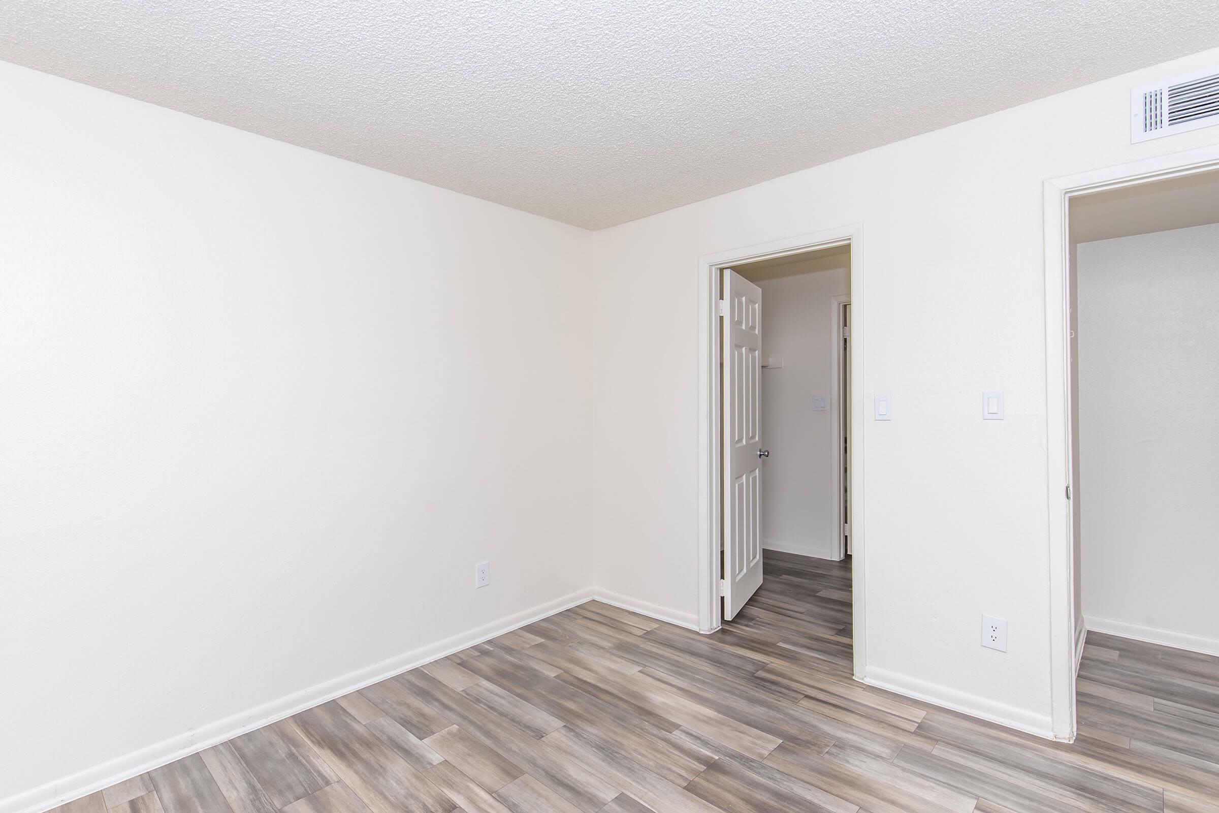 Empty room with light-colored walls and a textured ceiling. The floor features laminate planks in shades of brown. There are two doorways, one leading to another room and the other partially open. Natural light illuminates the space, creating a bright and airy atmosphere.