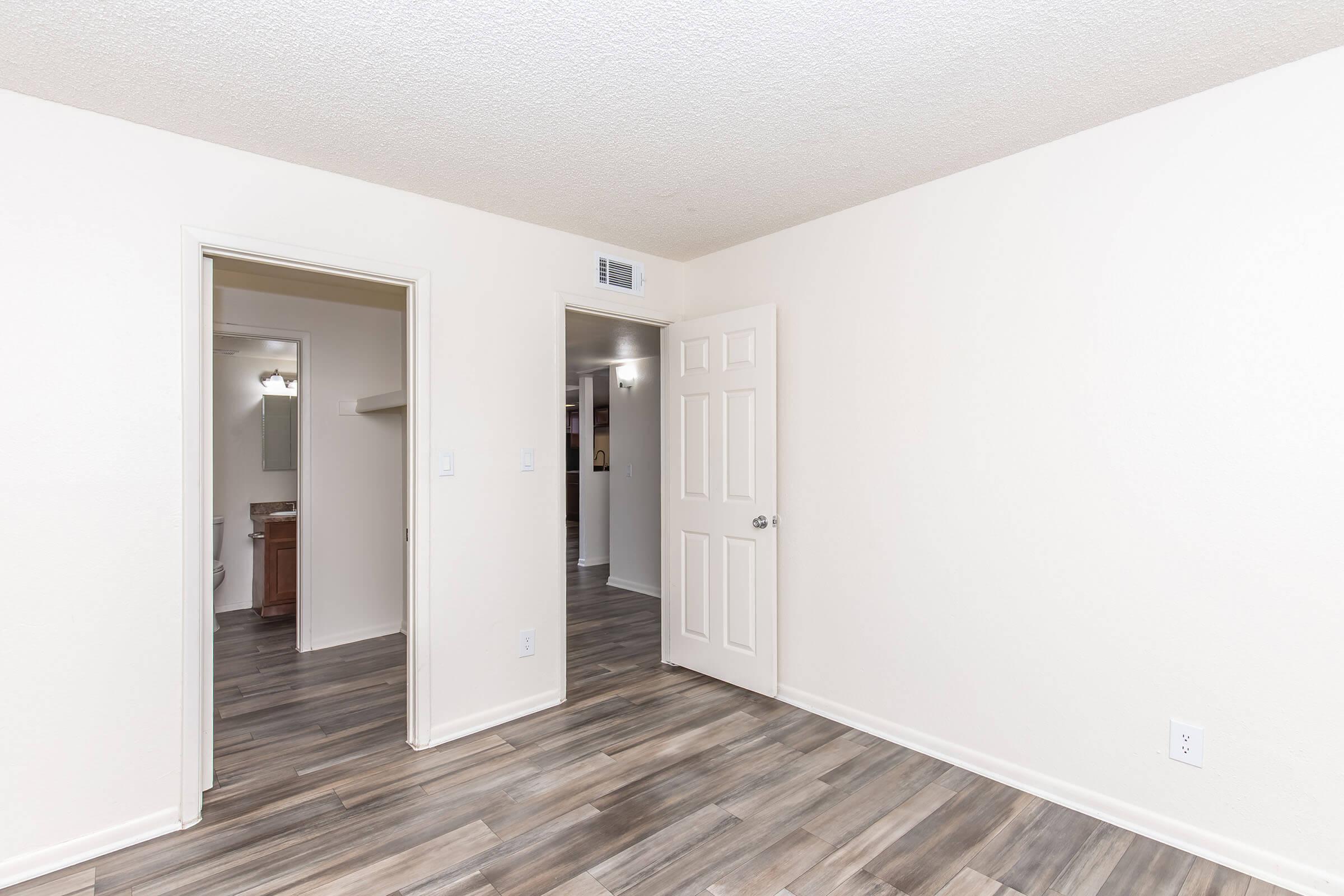 Interior view of a light-colored room with a textured ceiling and wooden flooring. Two doorways lead to adjacent spaces, one potentially a bathroom or closet. The walls are painted in a neutral shade, creating a bright and open atmosphere. The room appears freshly painted and well-maintained.