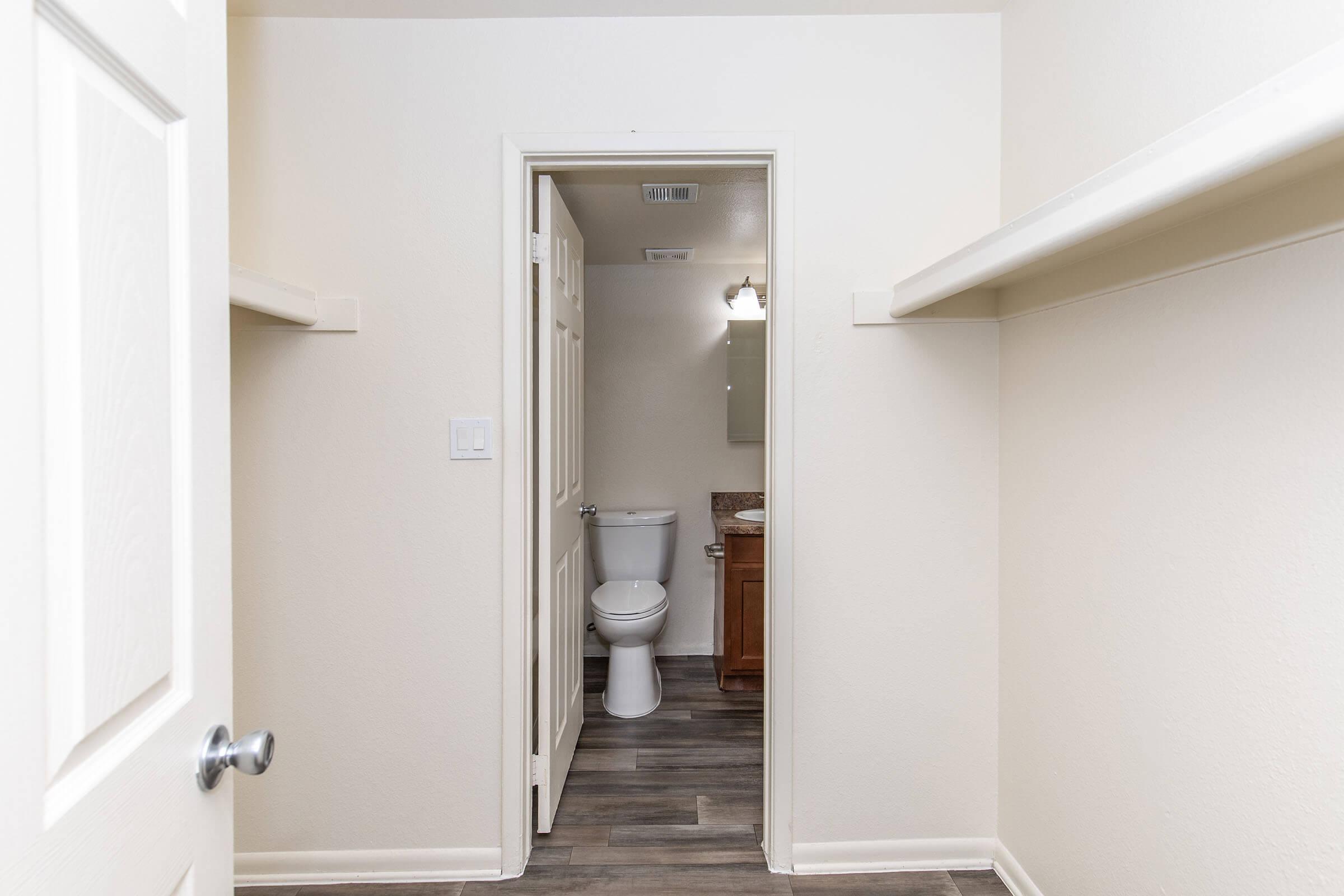 Interior view of a small bathroom with a toilet and sink, visible through an open doorway. Light beige walls and dark wood-look flooring create a neutral aesthetic. Two shelves are mounted on the walls, and a light fixture is visible above the mirror. The door to the room is ajar, leading to another space.