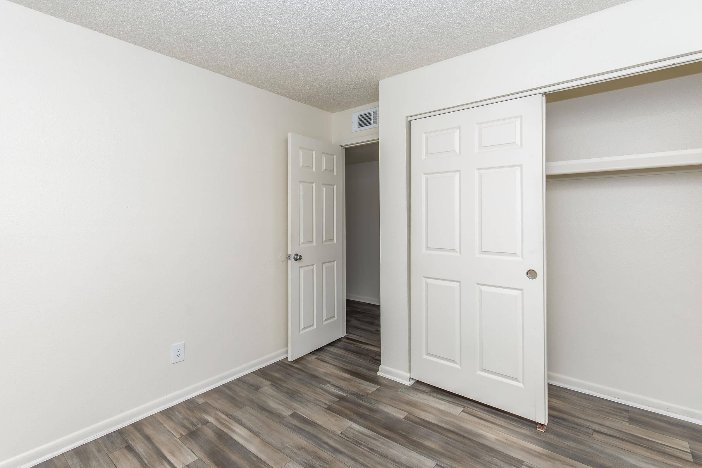Interior of a room featuring beige walls and wooden-style flooring. There are two closed white doors, one leading to a closet and the other to an adjacent space. Natural light fills the room, creating a bright and inviting atmosphere.