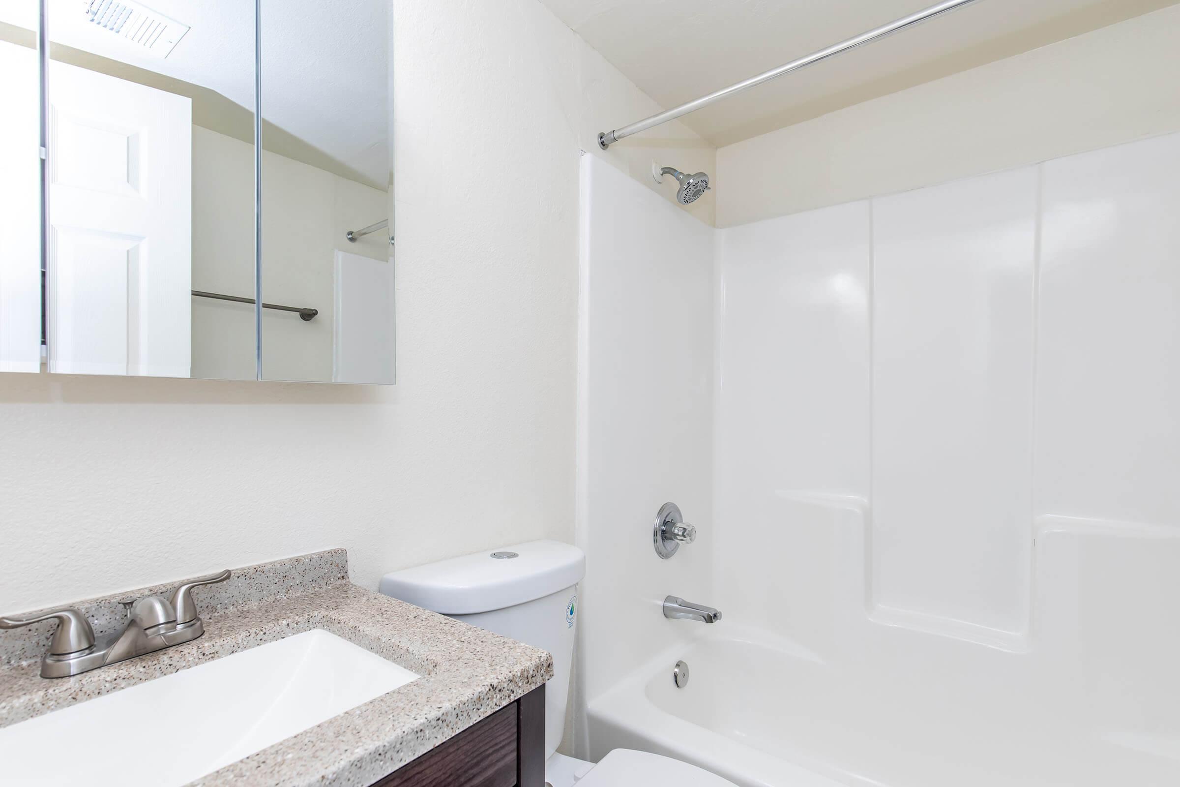 A clean bathroom featuring a white bathtub with a shower, a modern sink with a granite countertop, and a toilet. A large mirror is mounted on the wall above the sink, and the walls are painted a light color, creating a bright and open atmosphere.