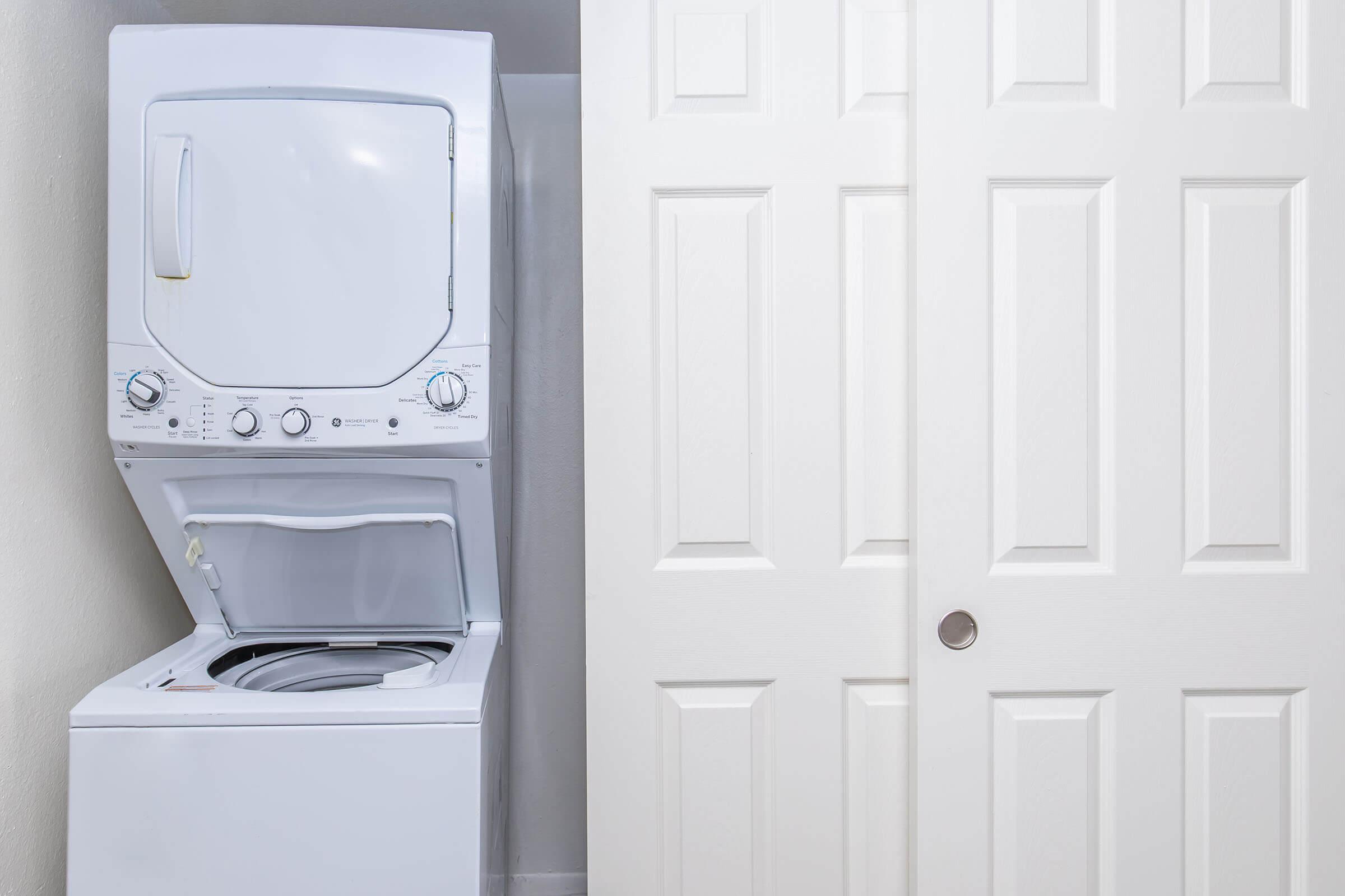 A stacked washer and dryer set in a laundry area, with the washer’s lid open. Adjacent to it is a set of white doors that appear to be closet doors, featuring a simple panel design. The room has neutral-colored walls and a clean, organized appearance.
