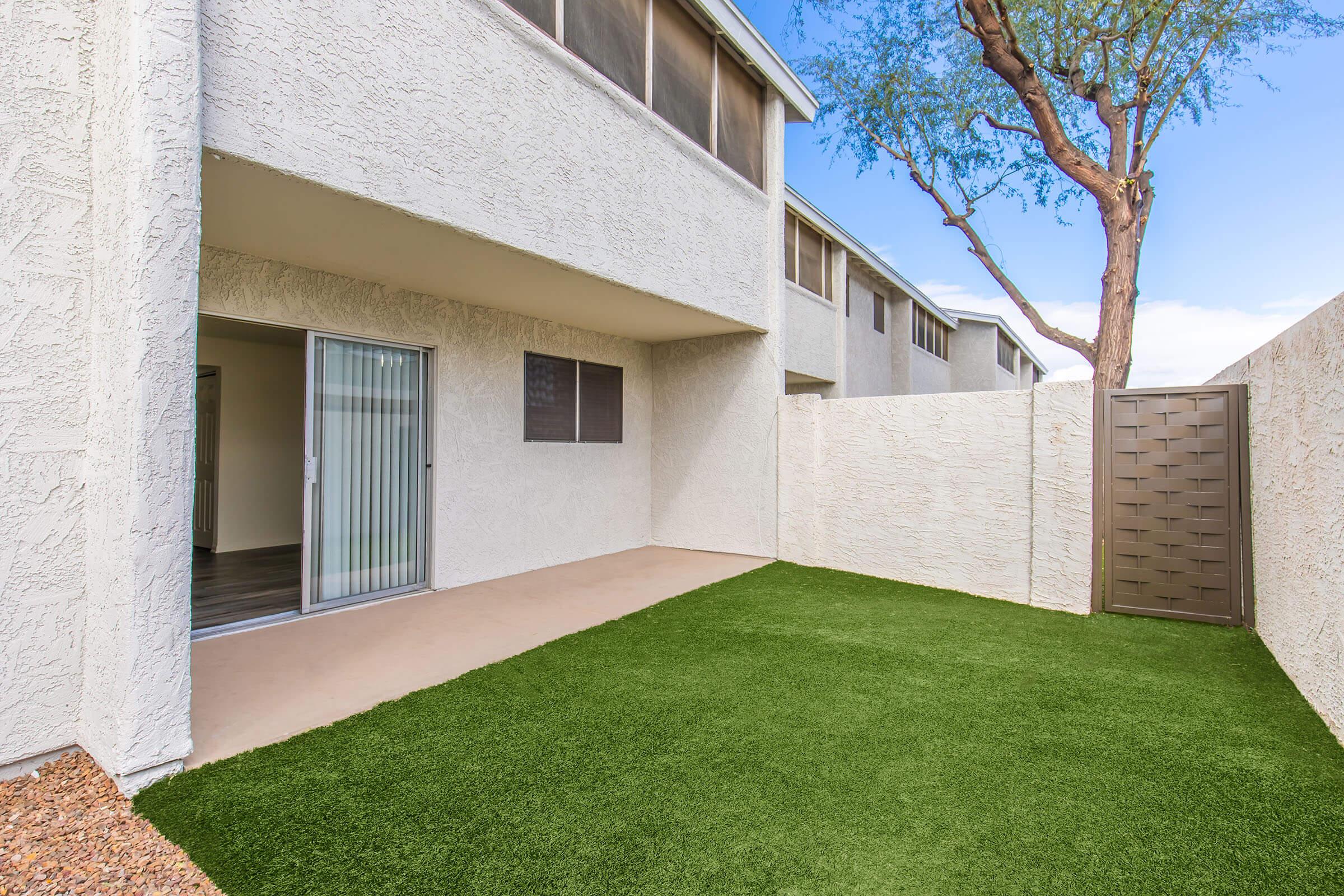 A small outdoor space featuring artificial grass, a concrete patio, and a tall wall for privacy. There's a sliding glass door leading indoors, and a decorative metal gate against one wall. Sparse landscaping includes a tree in the background and a clear blue sky above.