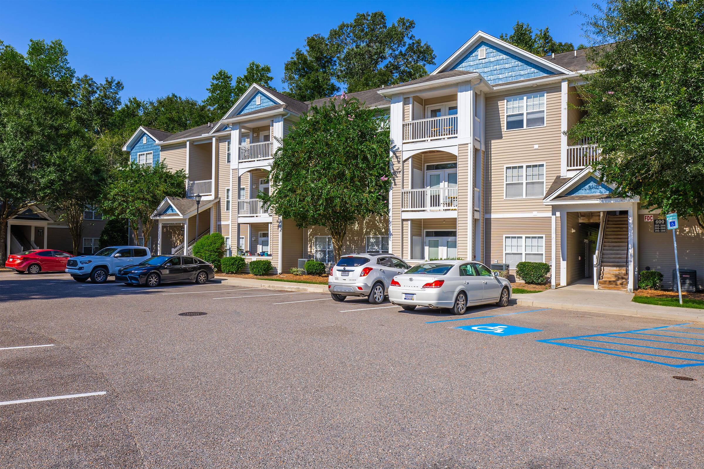 A well-maintained apartment complex with multiple buildings featuring balconies. The parking area in front has several cars parked, including a red vehicle and a white sedan. Lush green trees and shrubs surround the buildings, creating a pleasant and inviting atmosphere. Clear blue skies above.