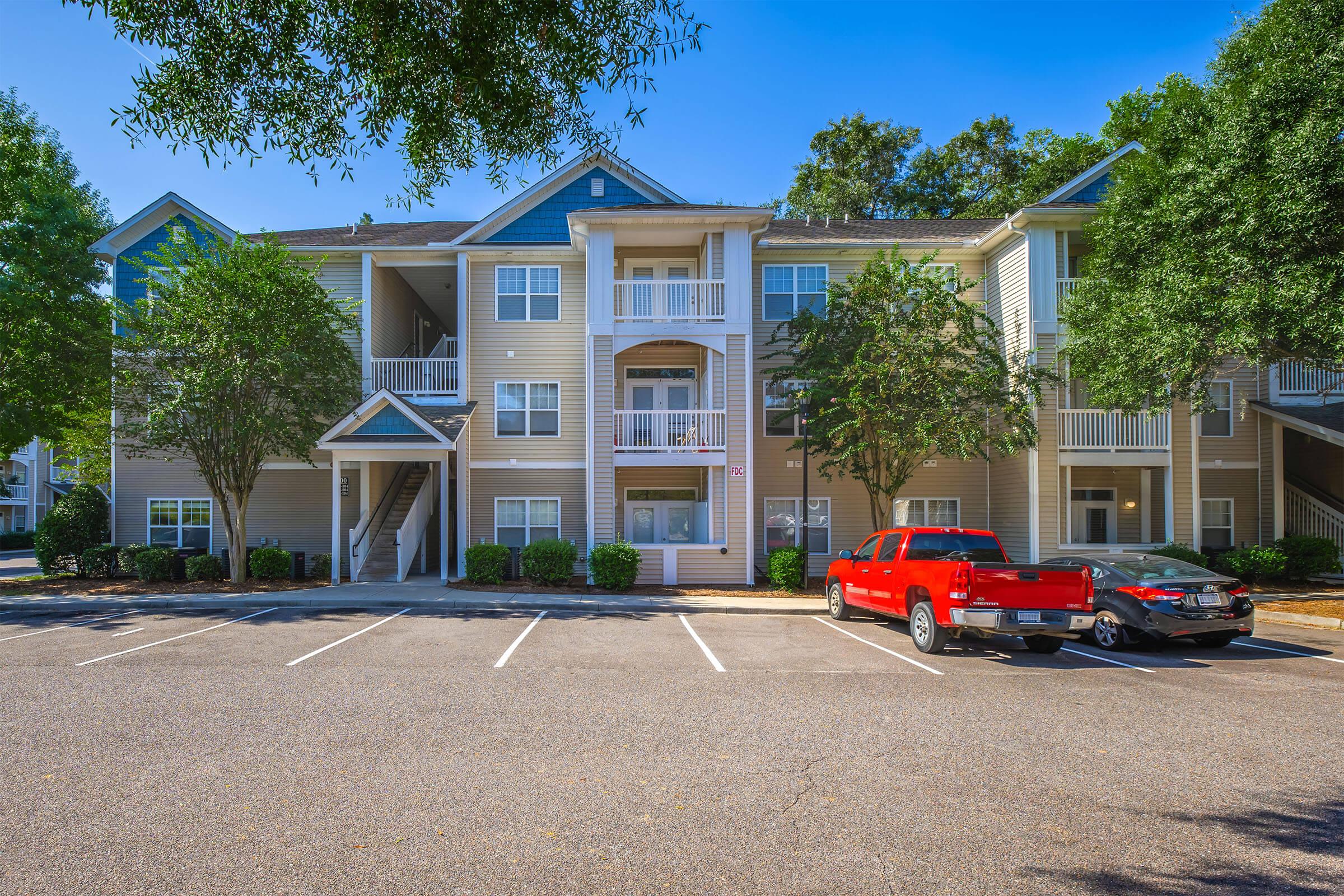 Modern apartment building with multiple balconies and staircases, surrounded by well-maintained landscaping and trees. A red pickup truck is parked in front, with additional vehicles in the parking area. The sky is clear and blue, indicating a sunny day.