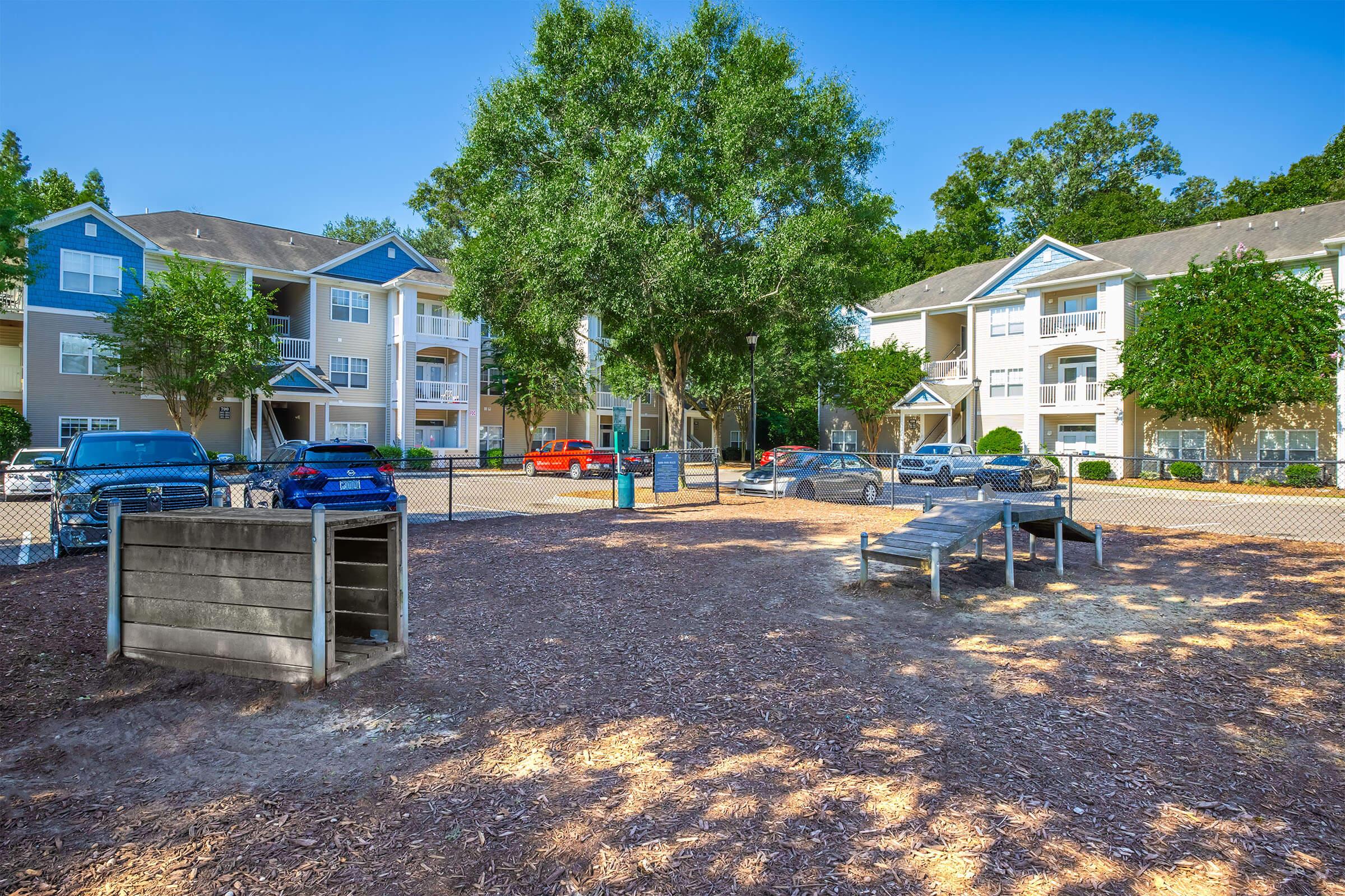 A playground area surrounded by apartment buildings, featuring a wooden climbing structure and a slide. The ground is covered with mulch, and there are trees providing shade. Several parked cars are visible in the background, indicating a residential community atmosphere. Bright blue skies add to the cheerful setting.