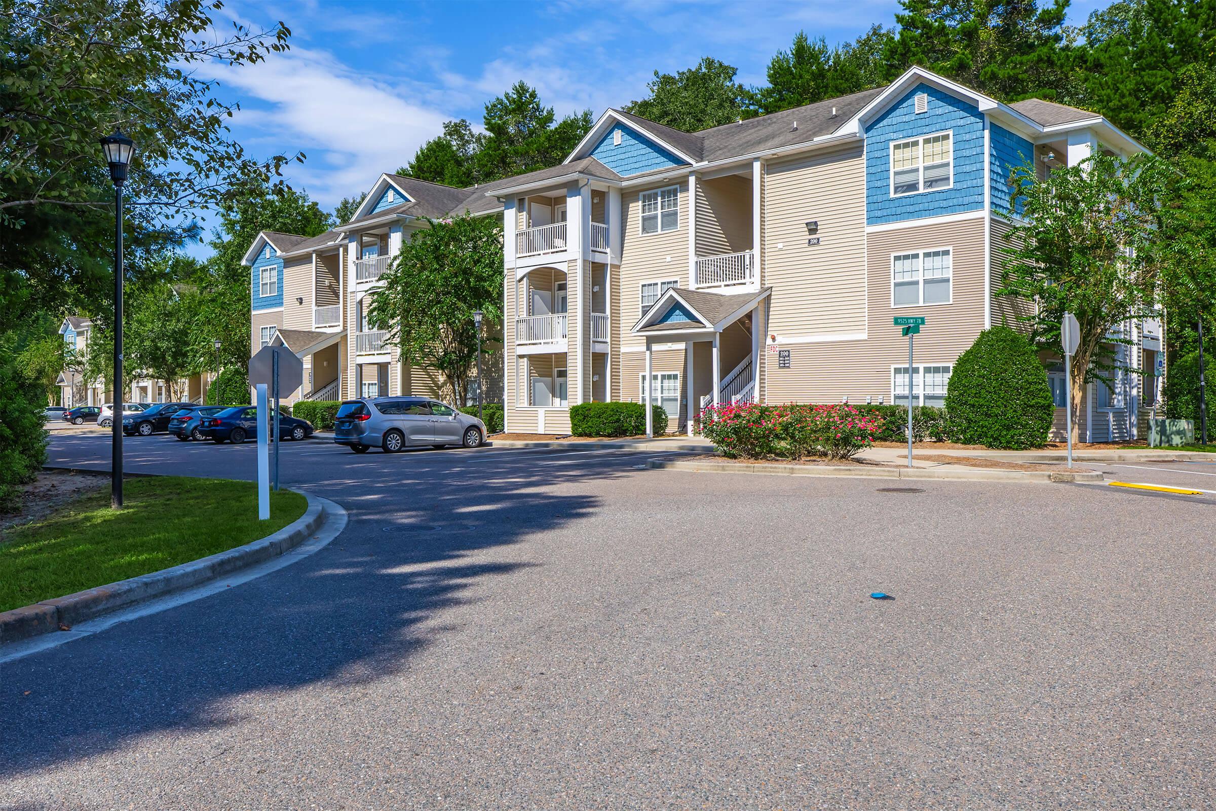 A residential apartment complex featuring three buildings with light blue and beige siding. The scene includes a well-maintained parking lot, landscaped greenery, and a clear blue sky. There are several parked cars and a street sign visible by the entrance to the complex.