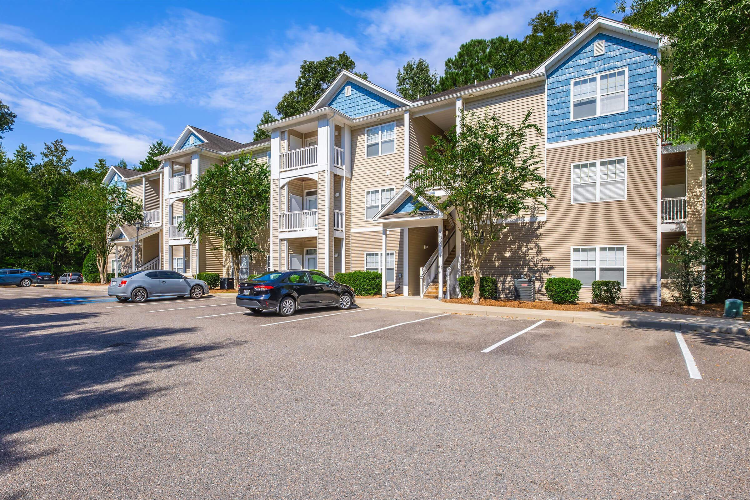 A three-story apartment building with light tan siding and blue accents. Trees and shrubs are planted near the entrance. There are several parking spaces in front, with two cars parked. The sky is clear and blue, indicating a sunny day.