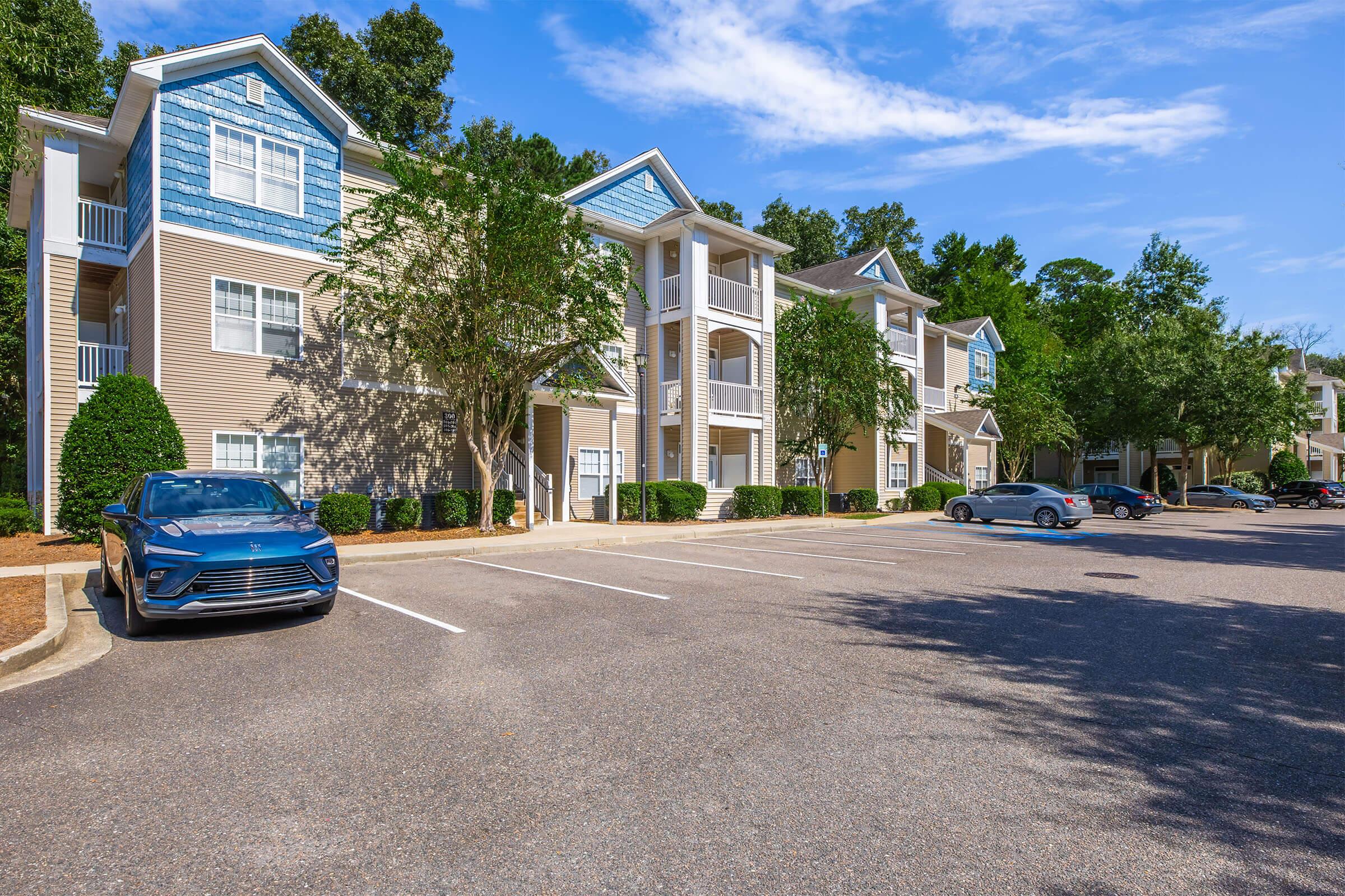 A view of an apartment complex featuring two-story buildings with blue accents. The scene includes a partially filled parking lot with several cars parked, surrounded by greenery and trees under a clear blue sky. The architecture is modern and well-maintained.
