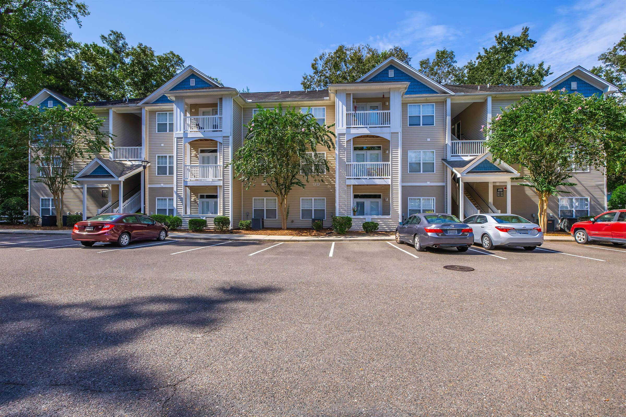 A three-story apartment building with a tan exterior and blue accents, surrounded by green trees and shrubs. Several parked cars are in the foreground, and the sky is clear with scattered clouds. Balconies are visible on the upper floors of the building.
