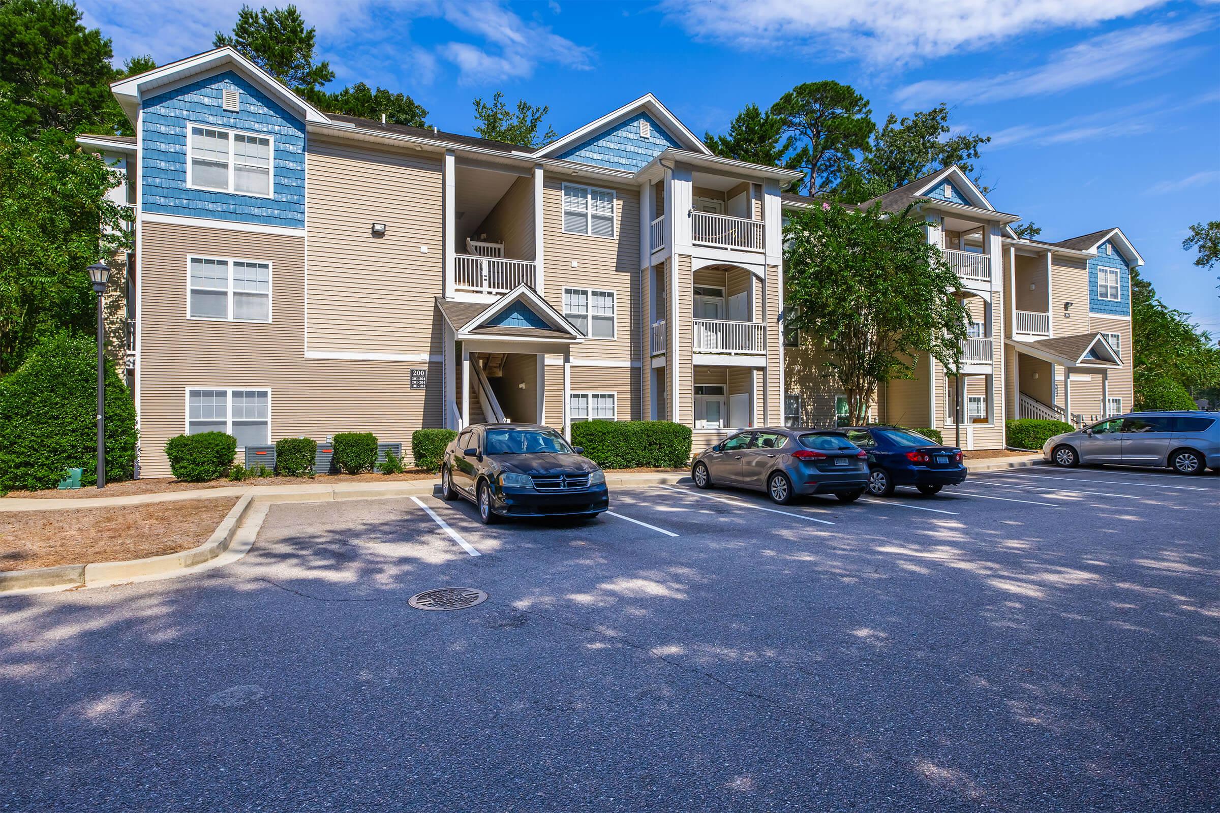 A multi-story apartment building with a light beige exterior and blue accents. The building features balconies and several windows. A parking lot in front has multiple vehicles parked, with green shrubs and trees surrounding the area under a bright blue sky with scattered clouds.