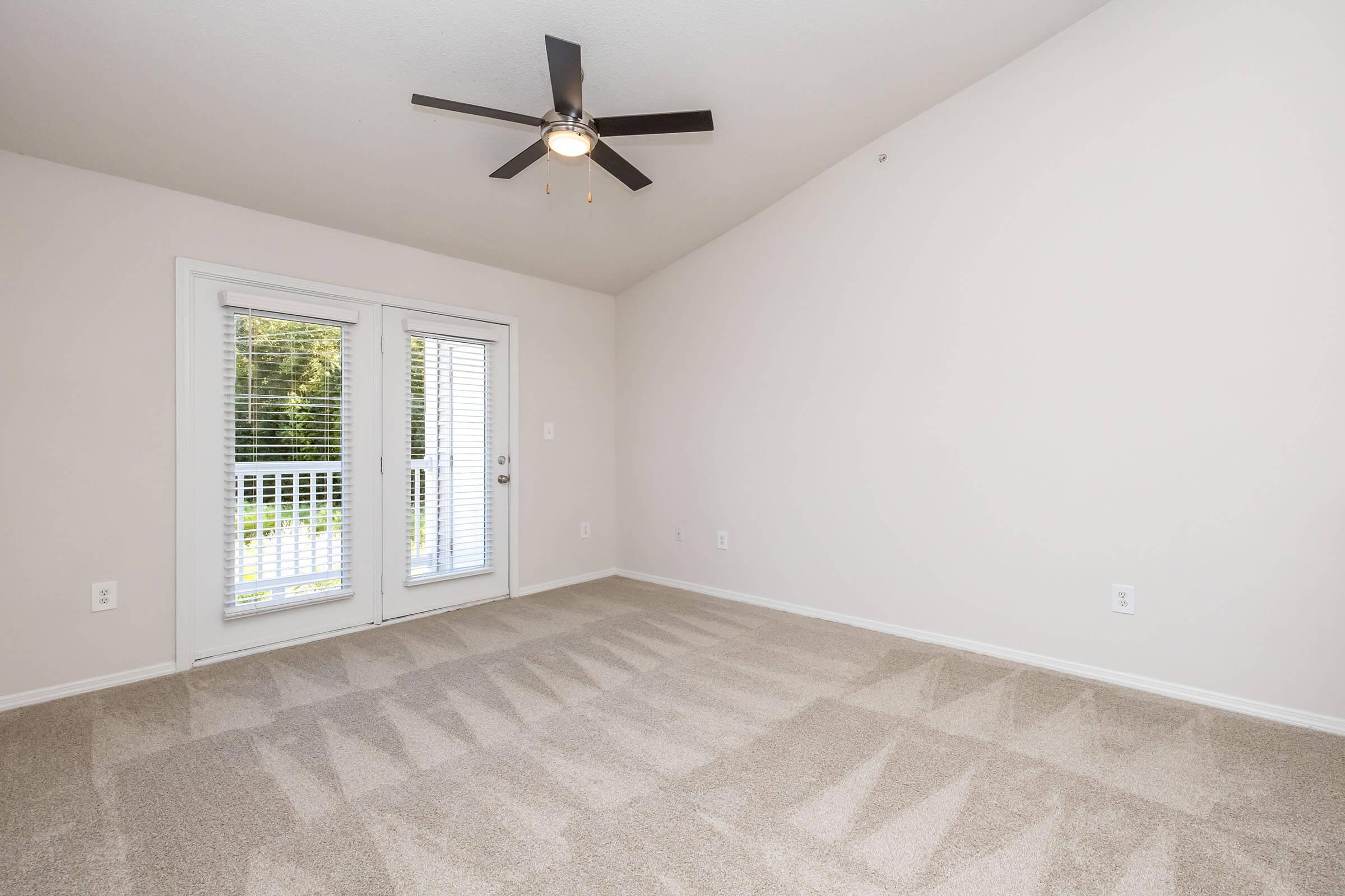 A spacious, empty room with light-colored walls and a ceiling fan. Carpeted floor with a noticeable pattern. There are large sliding glass doors leading to an outdoor area, adorned with white blinds. Natural light fills the space, creating a bright atmosphere.